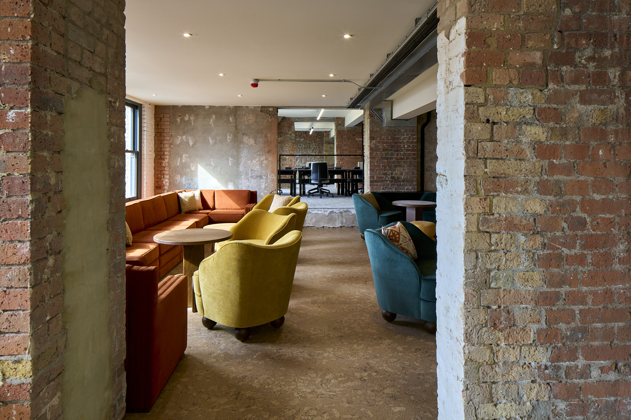 The Waterman lounge with rust corner sofa, yellow armchair and exposed plaster wall, next to raised workspace with height-adjustable desks