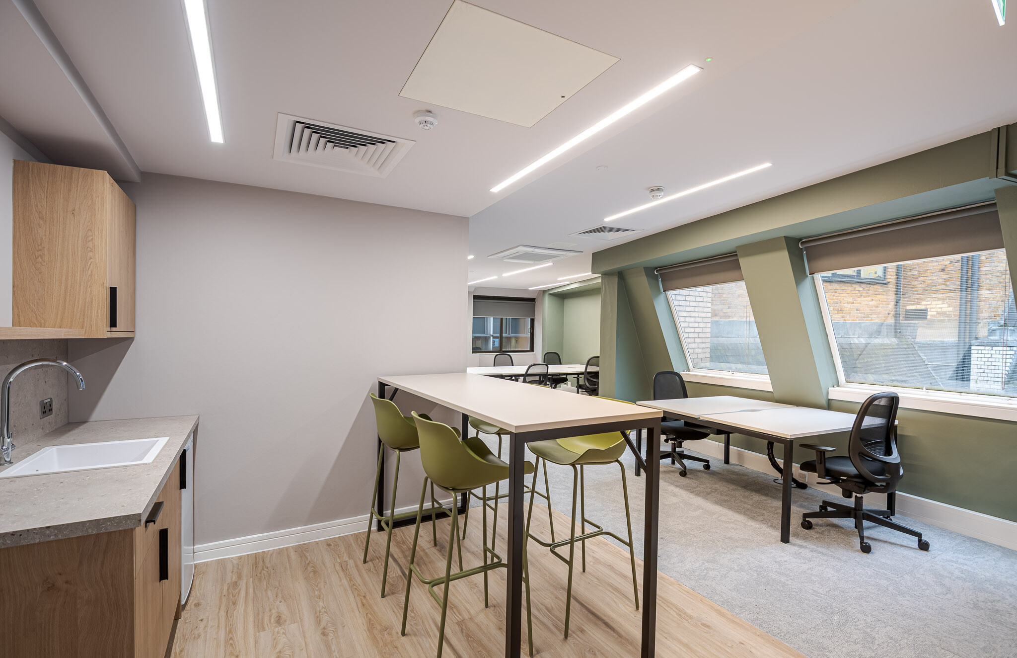 141 Fenchurch well-lit office kitchenette and workspace for Alchemy Asset Management, featuring a wooden teapoint, a high table with olive green stools, and large windows that bring in natural light. Workstations along the windows provide a bright and comfortable setting for focused work.