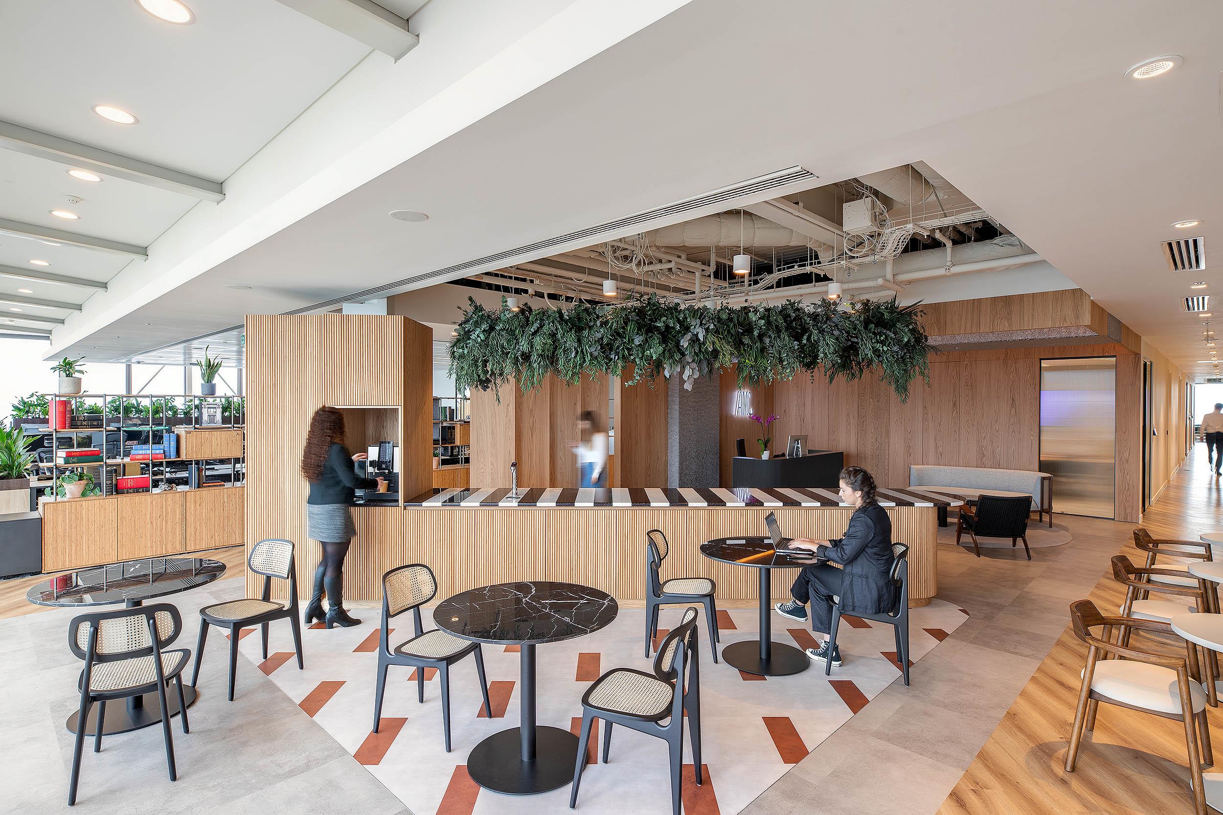 Furniture by Platfform for AMC’s café-style breakout space, featuring marble-topped tables, woven chairs and a timber coffee counter beneath hanging greenery.