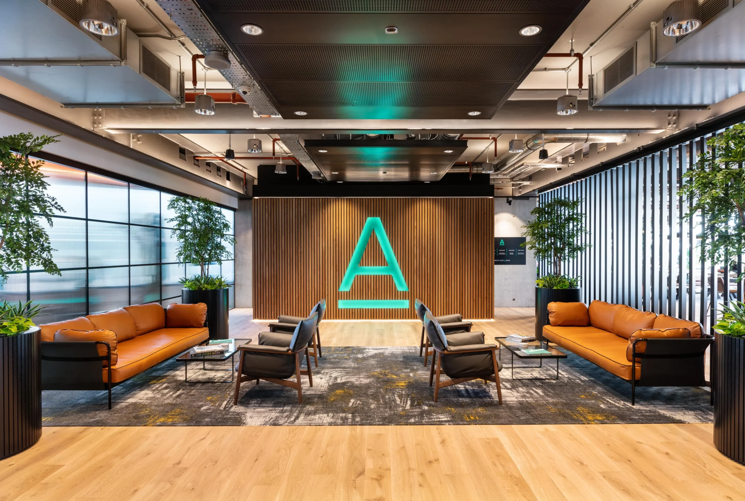 Reception area at Alpha FX, furnished by Platfform with warm leather sofas, contemporary armchairs, and biophilic accents beneath a glowing A logo.