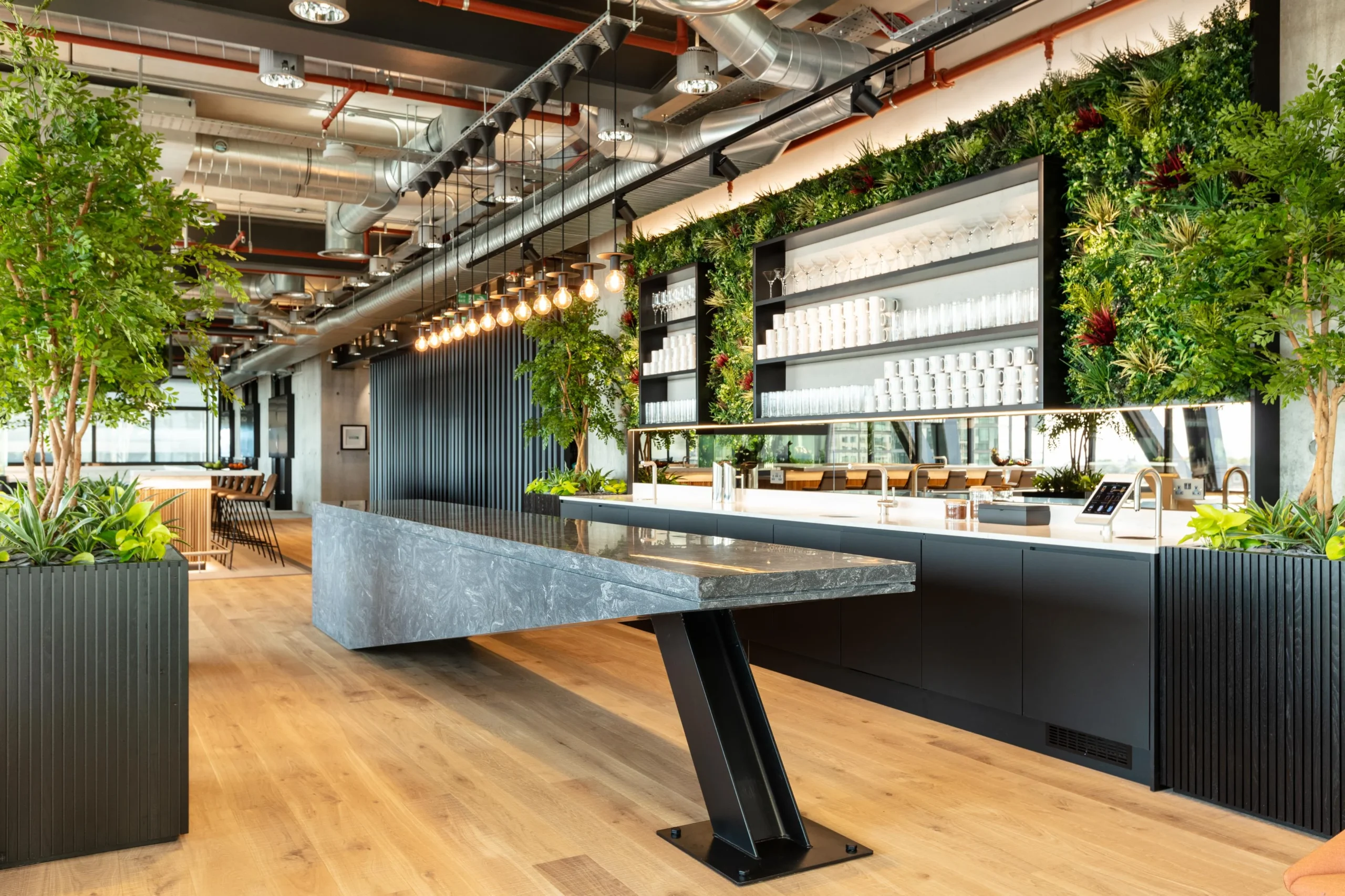 Alpha FX breakout space with bespoke furniture by Platfform, featuring a striking stone-effect table, open shelving, and lush planting integrated into the bar design.