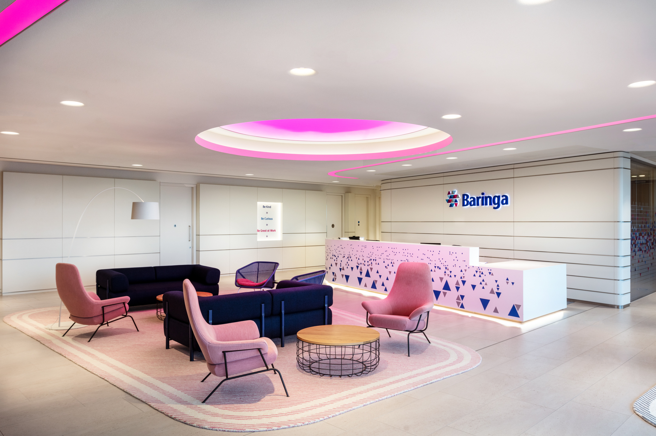 Reception area at Baringa with pink and navy lounge seating, geometric detailing on the desk, and a glowing magenta ceiling feature above.