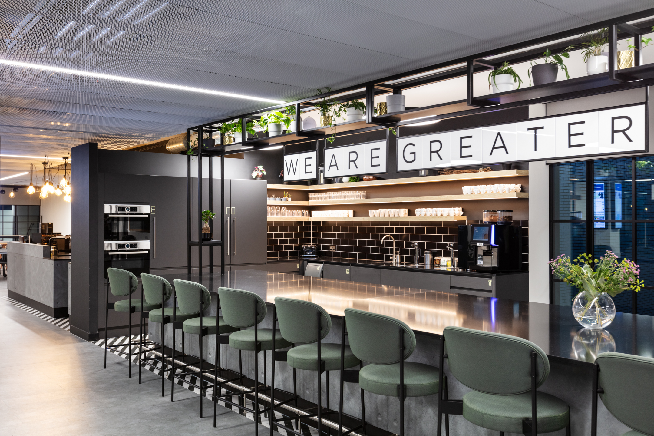 Kitchen and social hub at Checkout.com with black tiled splashback, open shelving, green bar stools and a bold ‘We Are Greater’ message overhead.