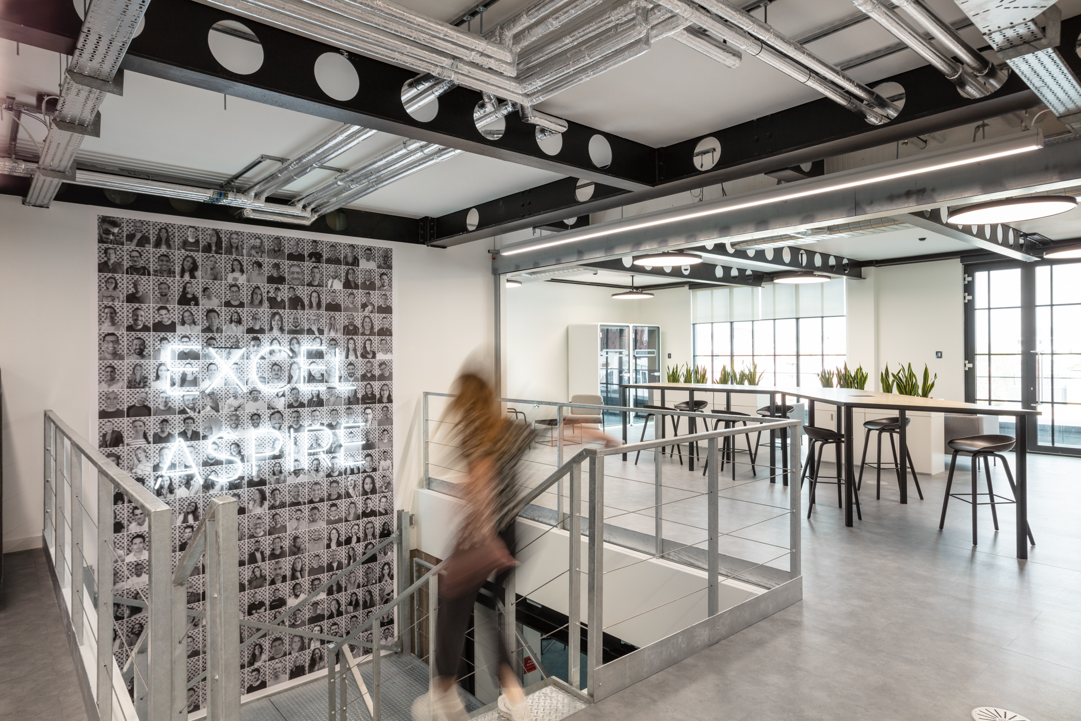 Upper floor at Checkout.com with industrial-style finishes, a high table workspace, and a neon-lit wall reading ‘Excel Aspire’ beside the stairwell.
