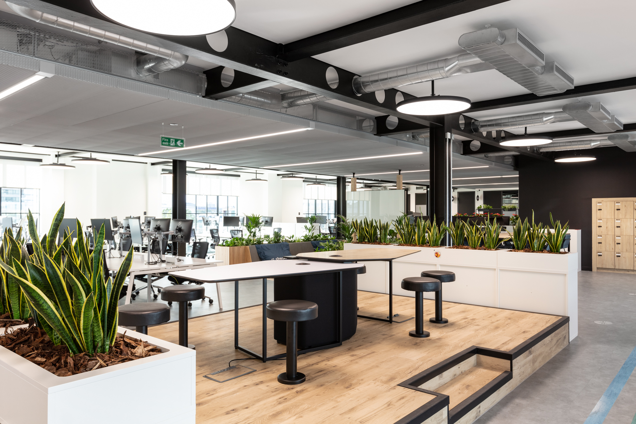 Collaborative working zone at Checkout.com with tiered flooring, bar-height tables, integrated planters and a backdrop of open desking.