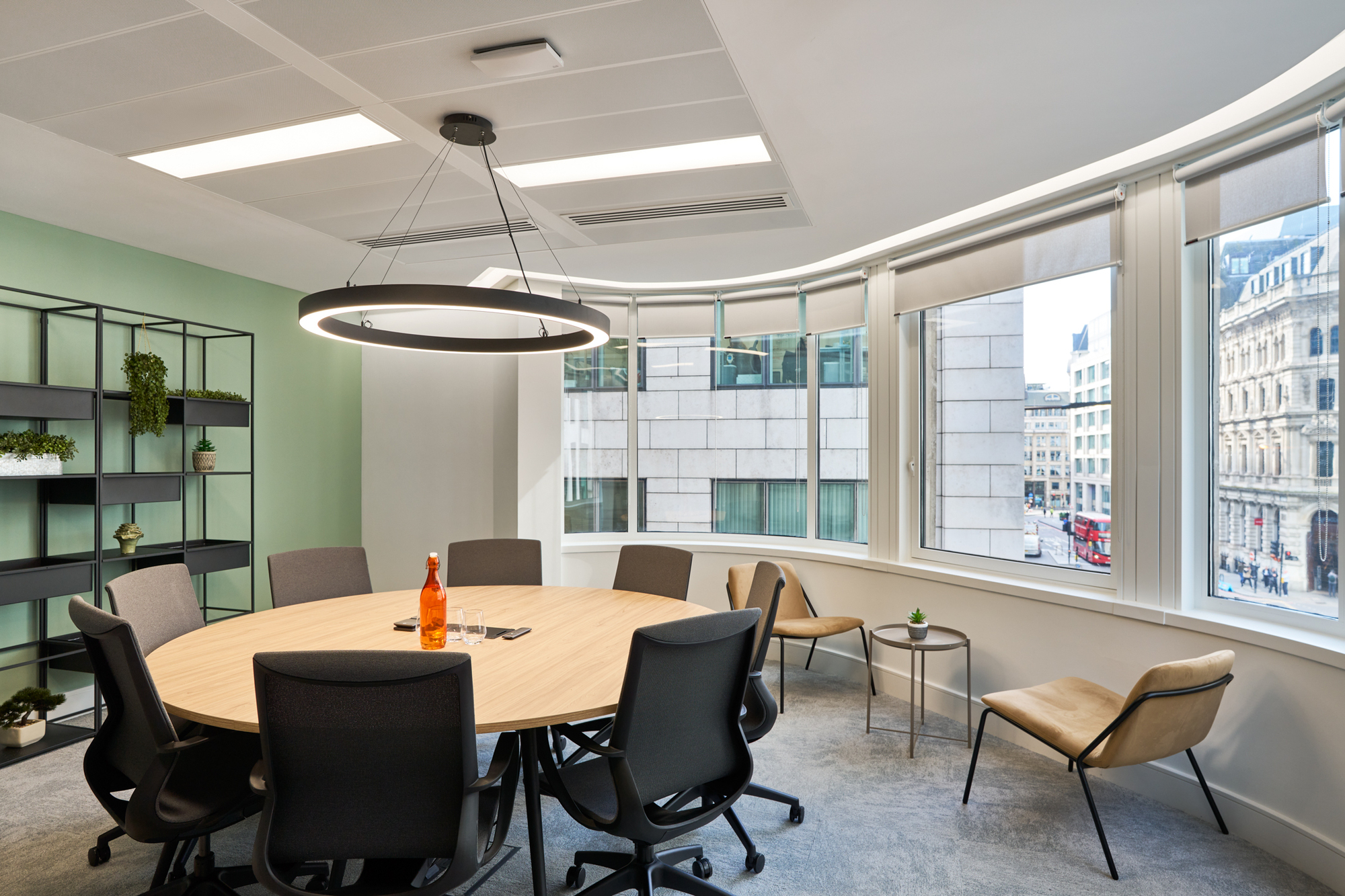 Round meeting room table at Gemserv’s workspace by Platfform, complemented by ergonomic chairs, floor-to-ceiling windows, and a modern circular light fixture.