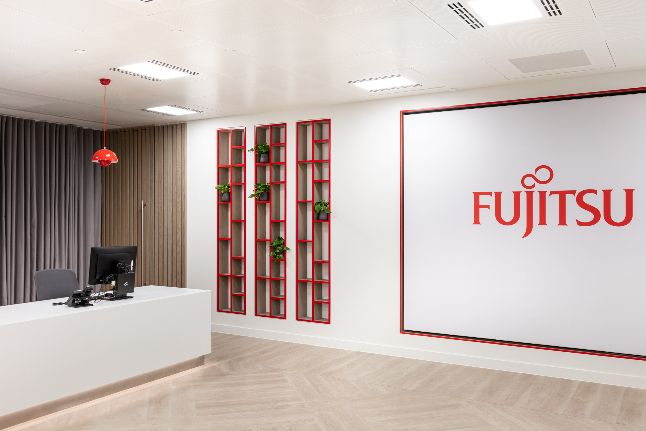 Reception area at Marylebone Fujitsu office featuring a sleek white desk, red pendant lighting and branded shelving with plants set against a statement logo wall.