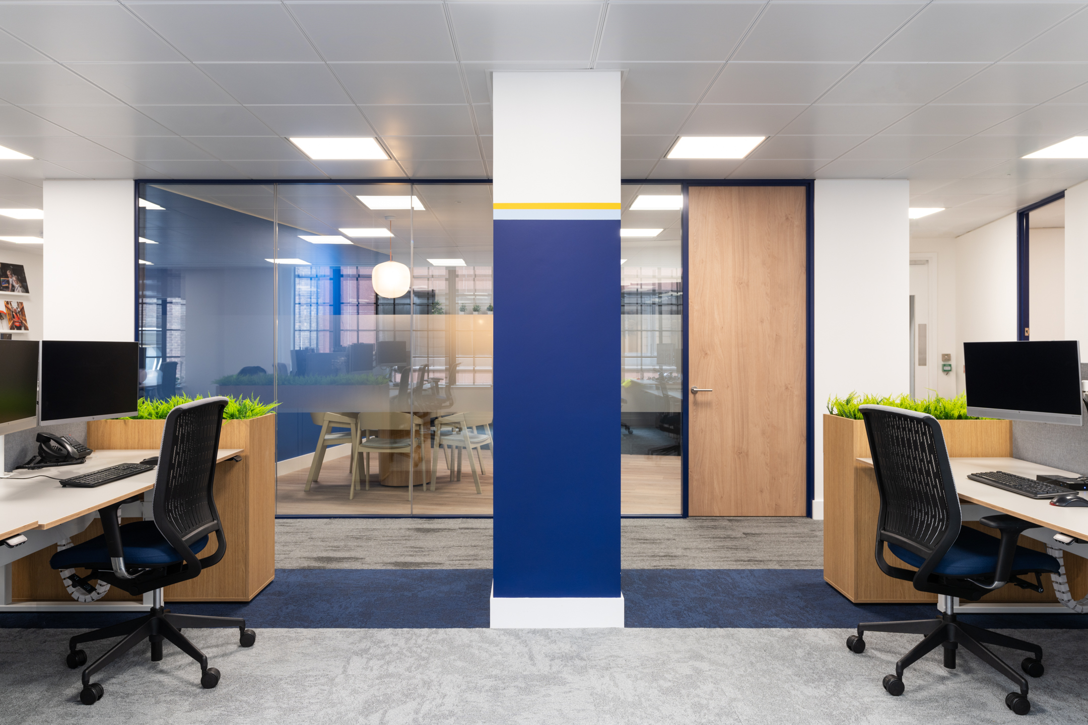 Modern workspace at the Port of London Authority office by Platfform, featuring desk partitions with greenery, a bold blue pillar, and a glass meeting room.