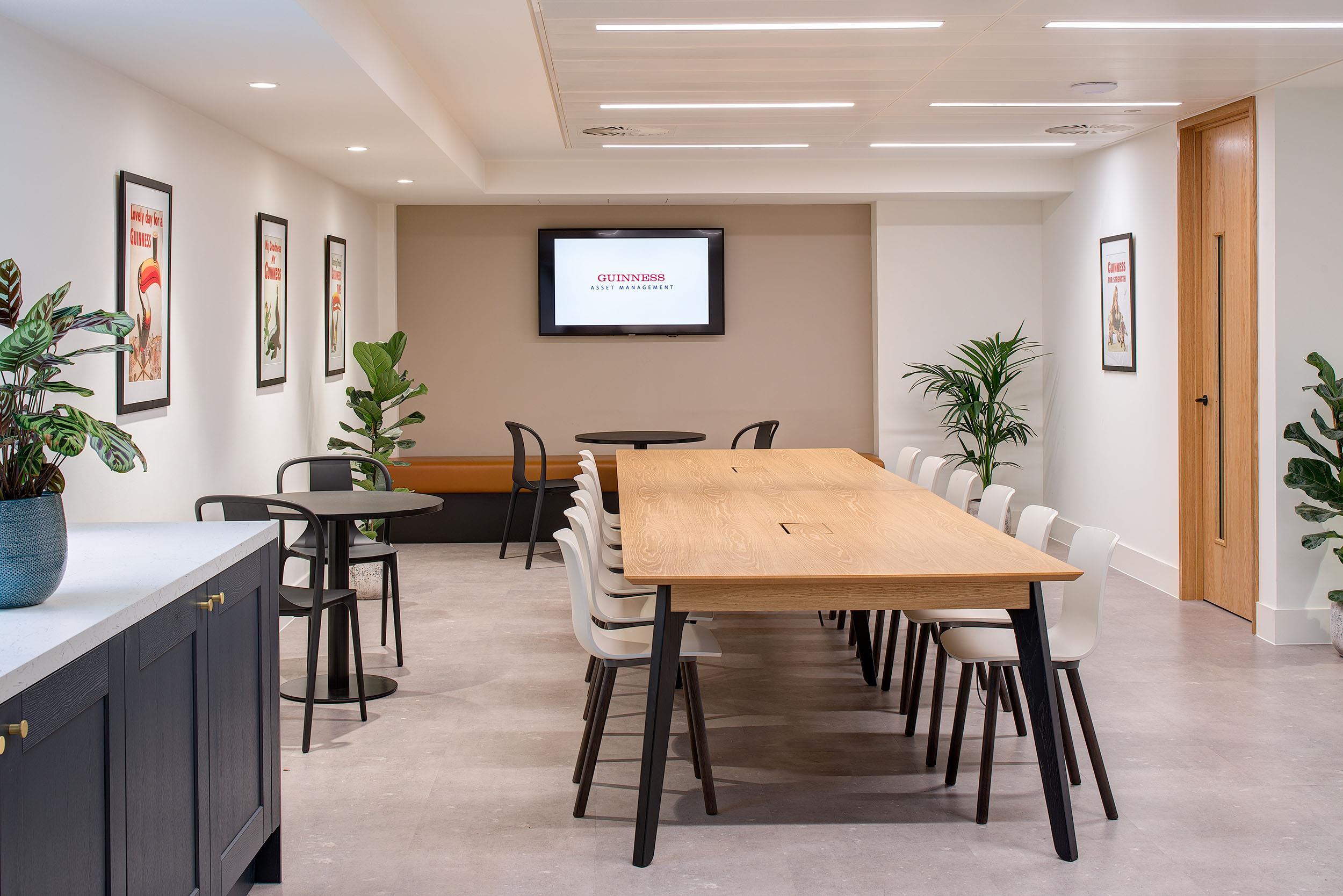 Breakout space at Guinness Asset Management furnished by Platfform, showing a long wooden table with white chairs, café-style seating, and a wall-mounted screen.