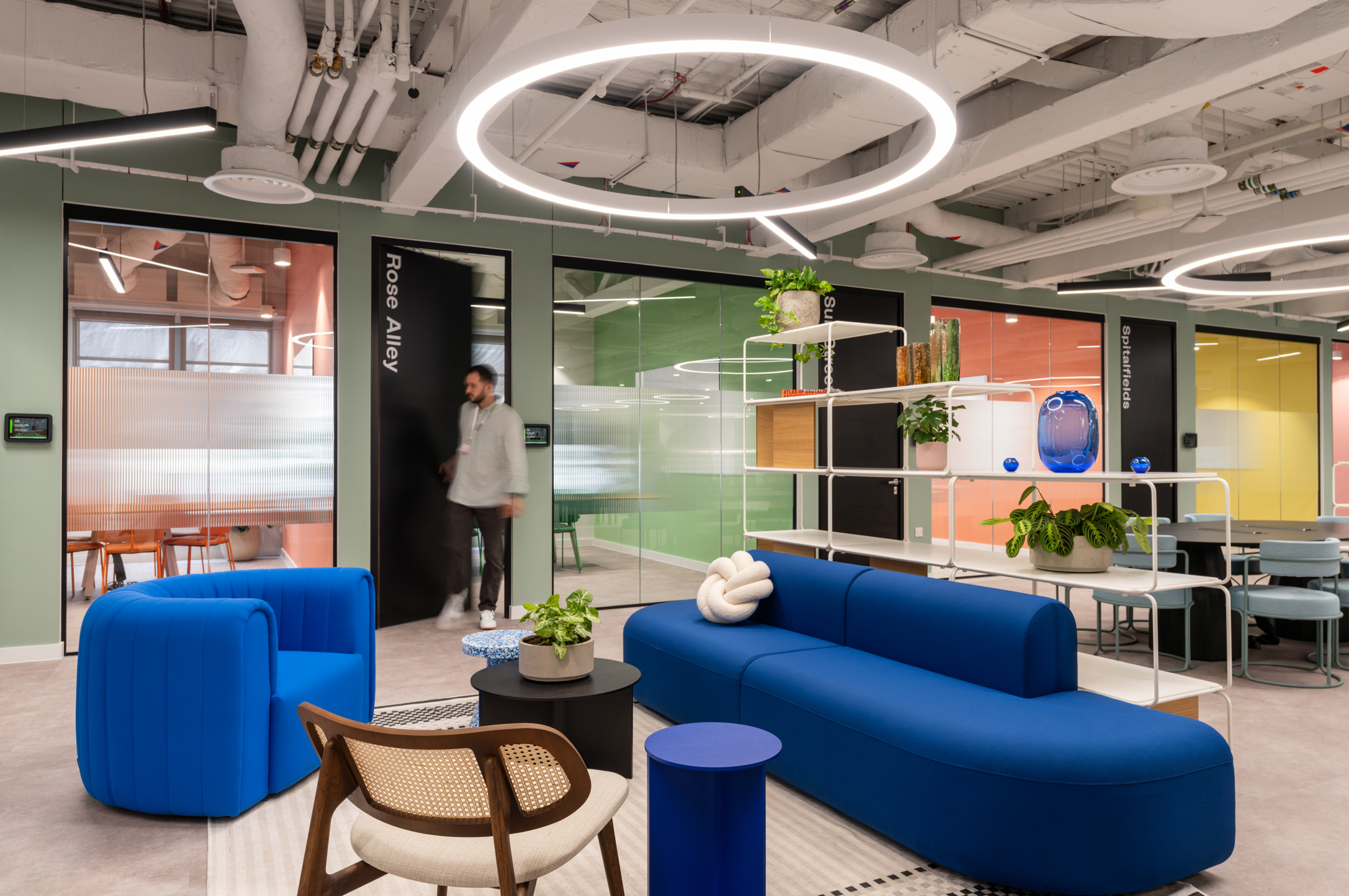 Breakout space at Huckletree, Bishopsgate, with bold blue lounge furniture, open shelving and a backdrop of colourful glass-fronted meeting rooms.