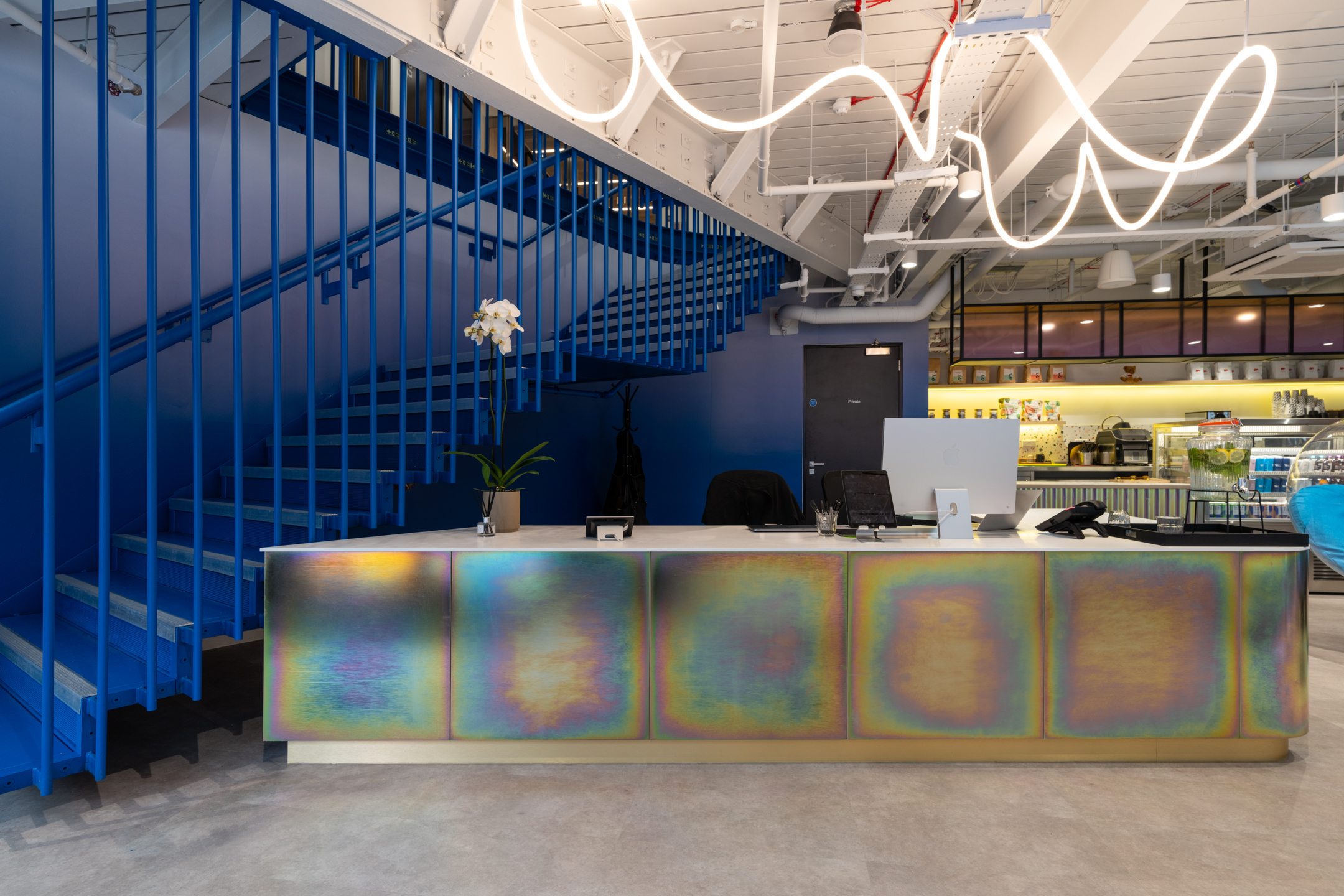 Reception at Huckletree, Bishopsgate, with a rainbow-iridescent desk, bold blue staircase and café backdrop lit by sculptural ceiling lighting.