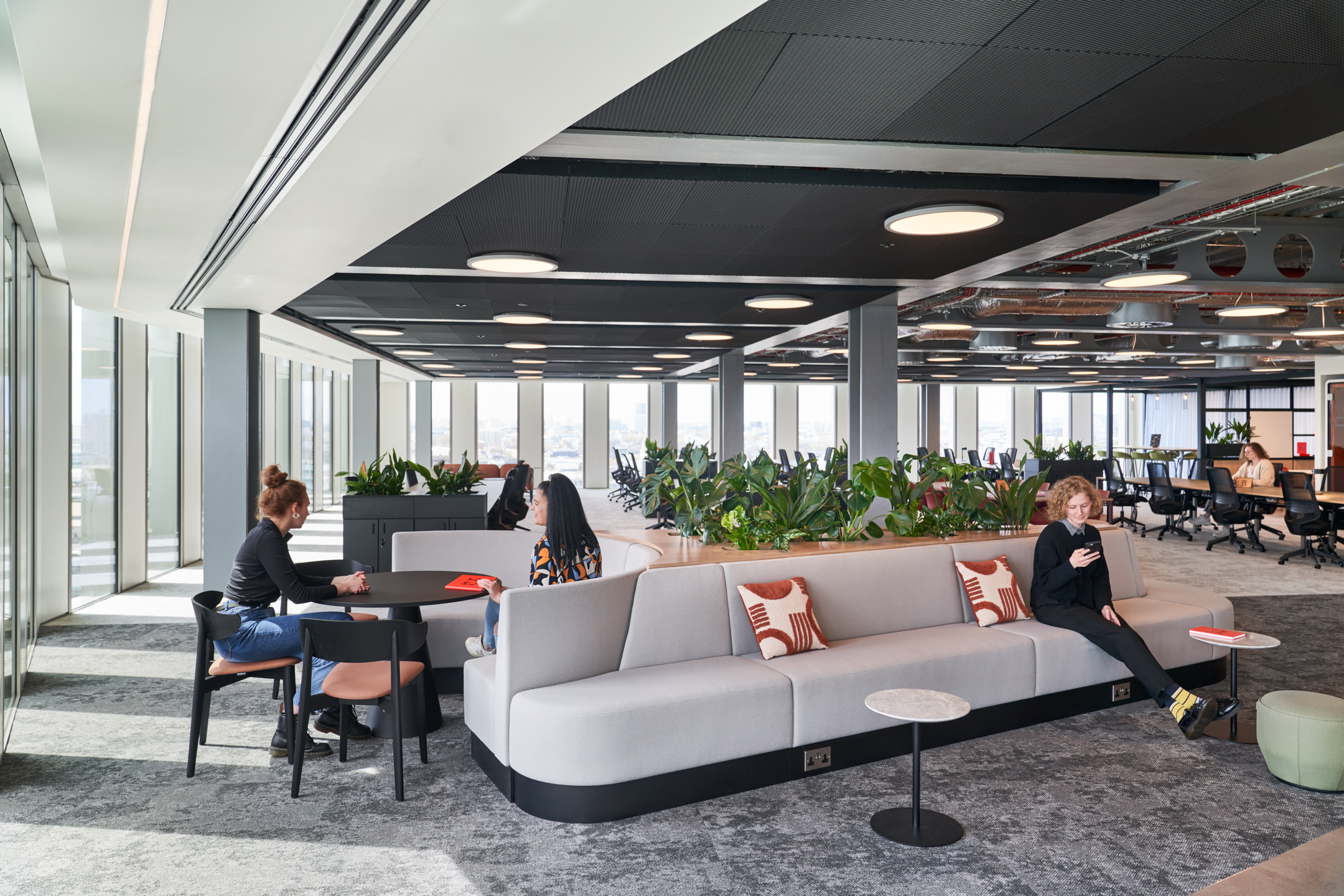 Flexible breakout area at HYLO with soft grey banquette seating, black café tables and layered planting, with furniture by Platfform.