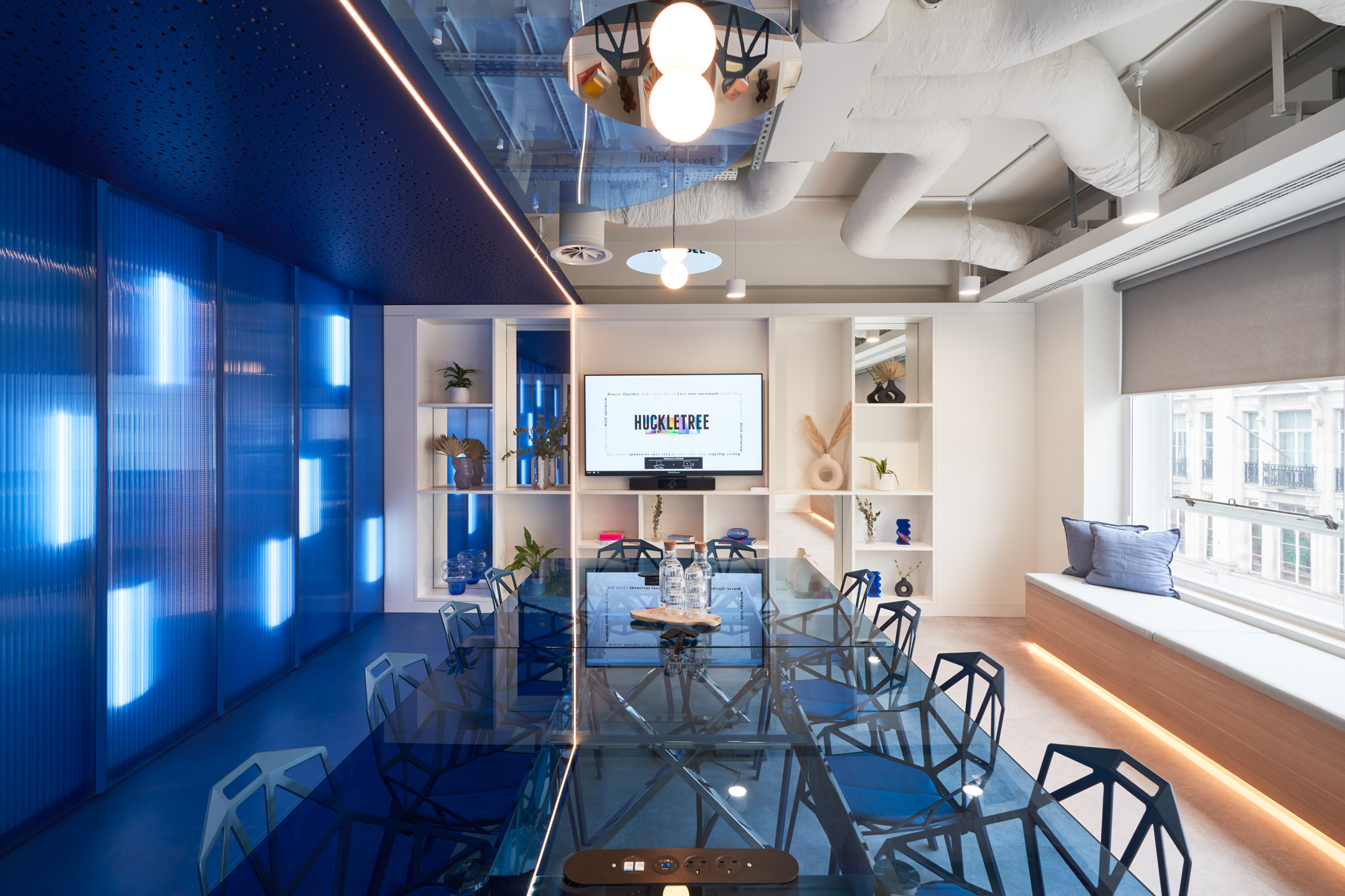 Glass meeting table and angular blue chairs styled by Platfform for Huckletree Oxford Street, set against a bold illuminated wall and vibrant shelving.