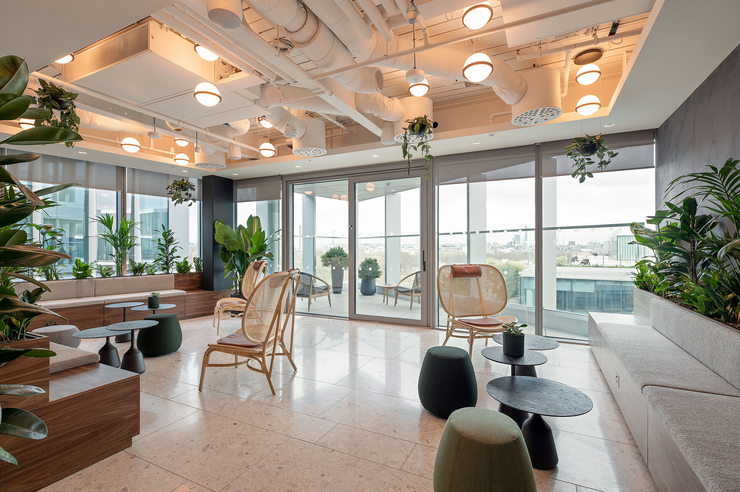 Bright indoor garden lounge at International Technology and Data Firm with rattan chairs, low tables and integrated planting beside a terrace entrance.