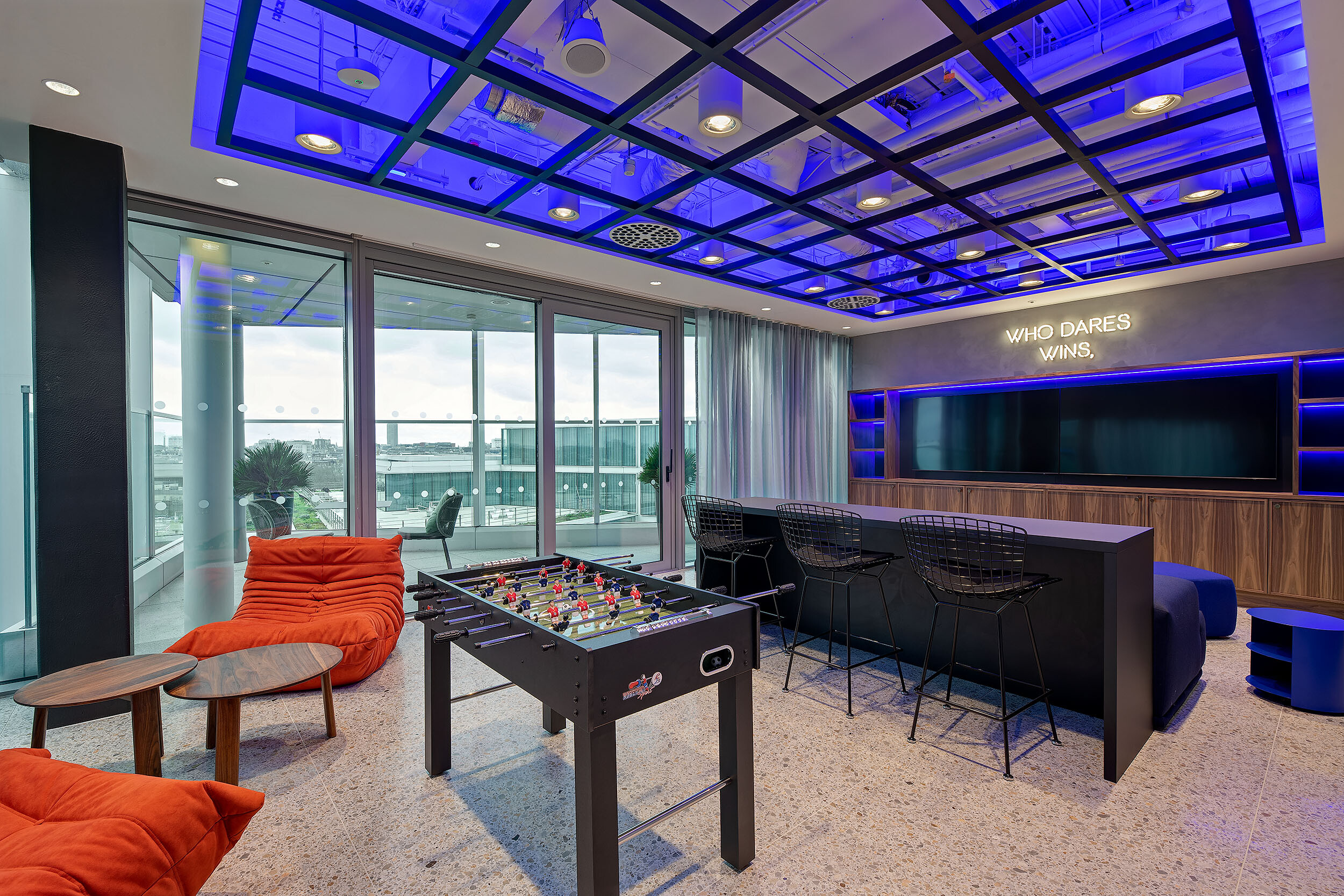 Gaming room at International Technology and Data Firm with lounge chairs, foosball table, bar seating and bold lighting for social downtime and team bonding.