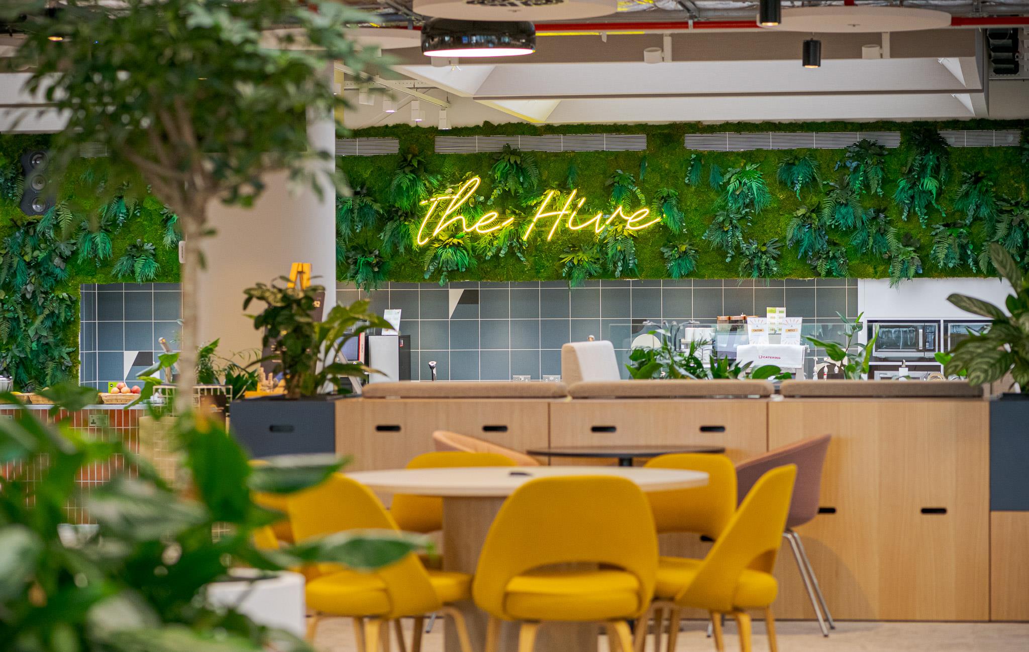 Vibrant breakout area at Landsec’s HQ, with yellow seating, softwood storage and a green living wall behind the service counter, lit by a neon ‘The Hive’ sign.