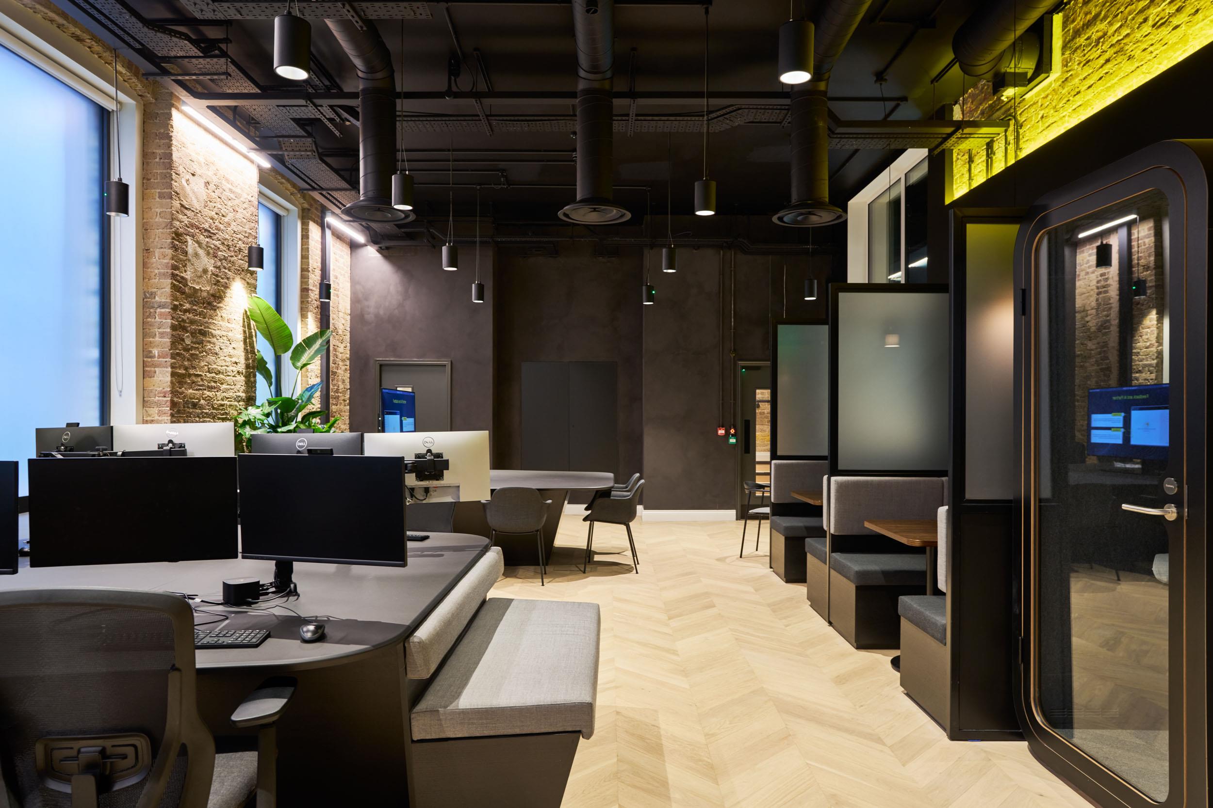 Workstations, meeting pods, and booth seating supplied for Lotus, styled within a dark interior palette and softened with planting and layered lighting.