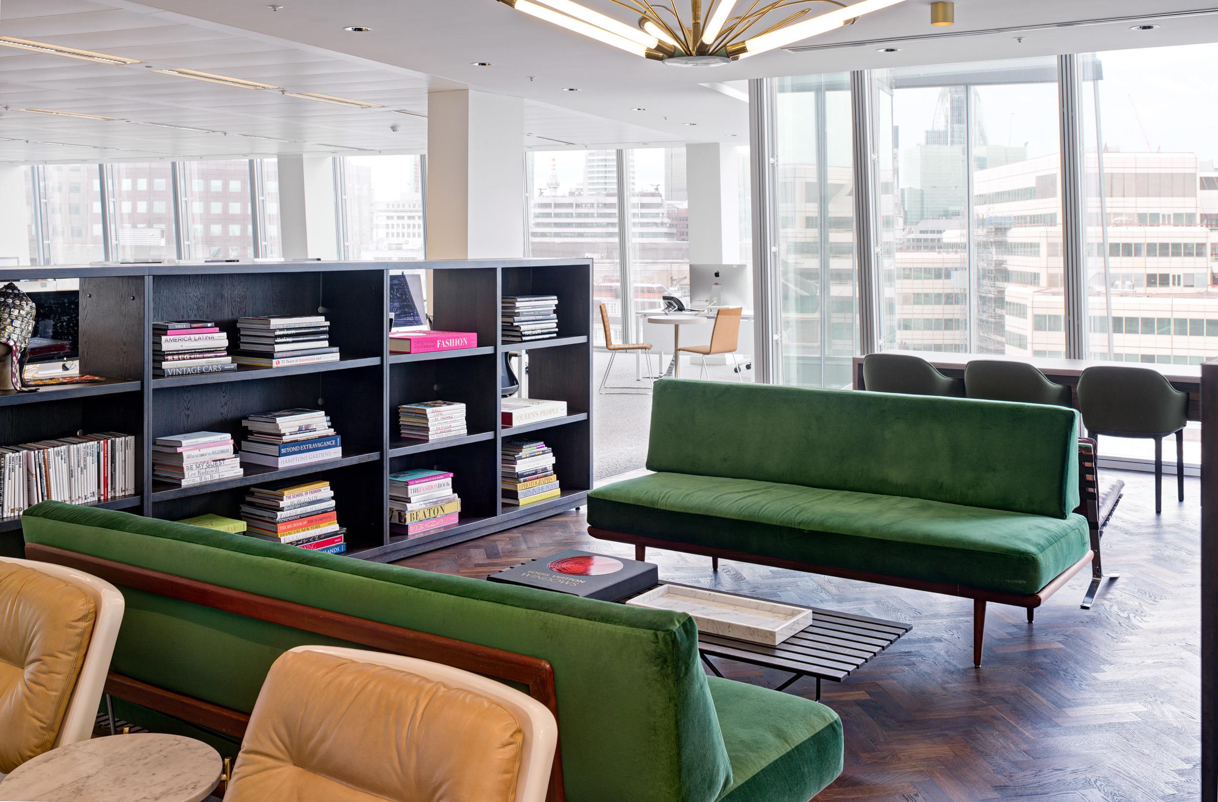 Lounge area at Matches Fashion, styled with green velvet sofas, tan leather armchairs, a black shelving unit filled with books, and city views through full-height glazing.