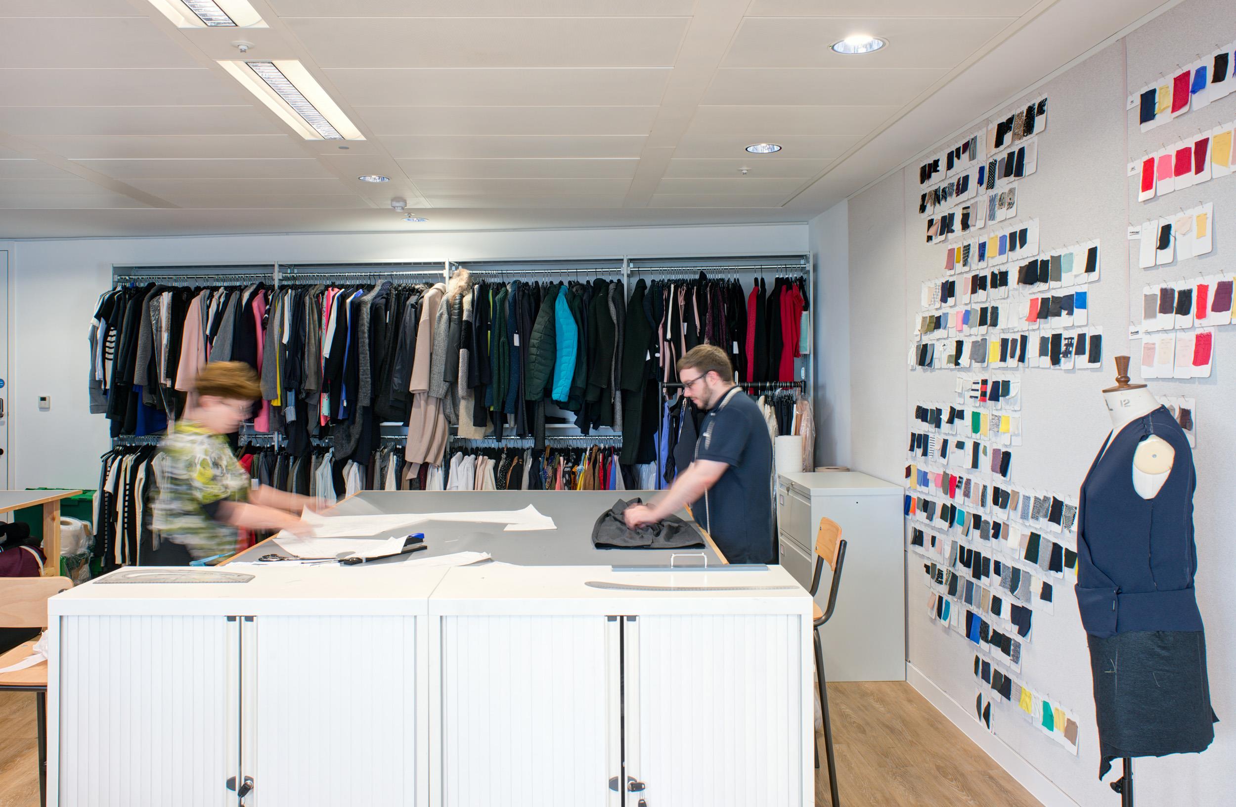 Fashion studio at Matches Fashion, with hanging garment rails, a central workbench, wall-mounted fabric swatches, and a mannequin dressed in a sample garment.