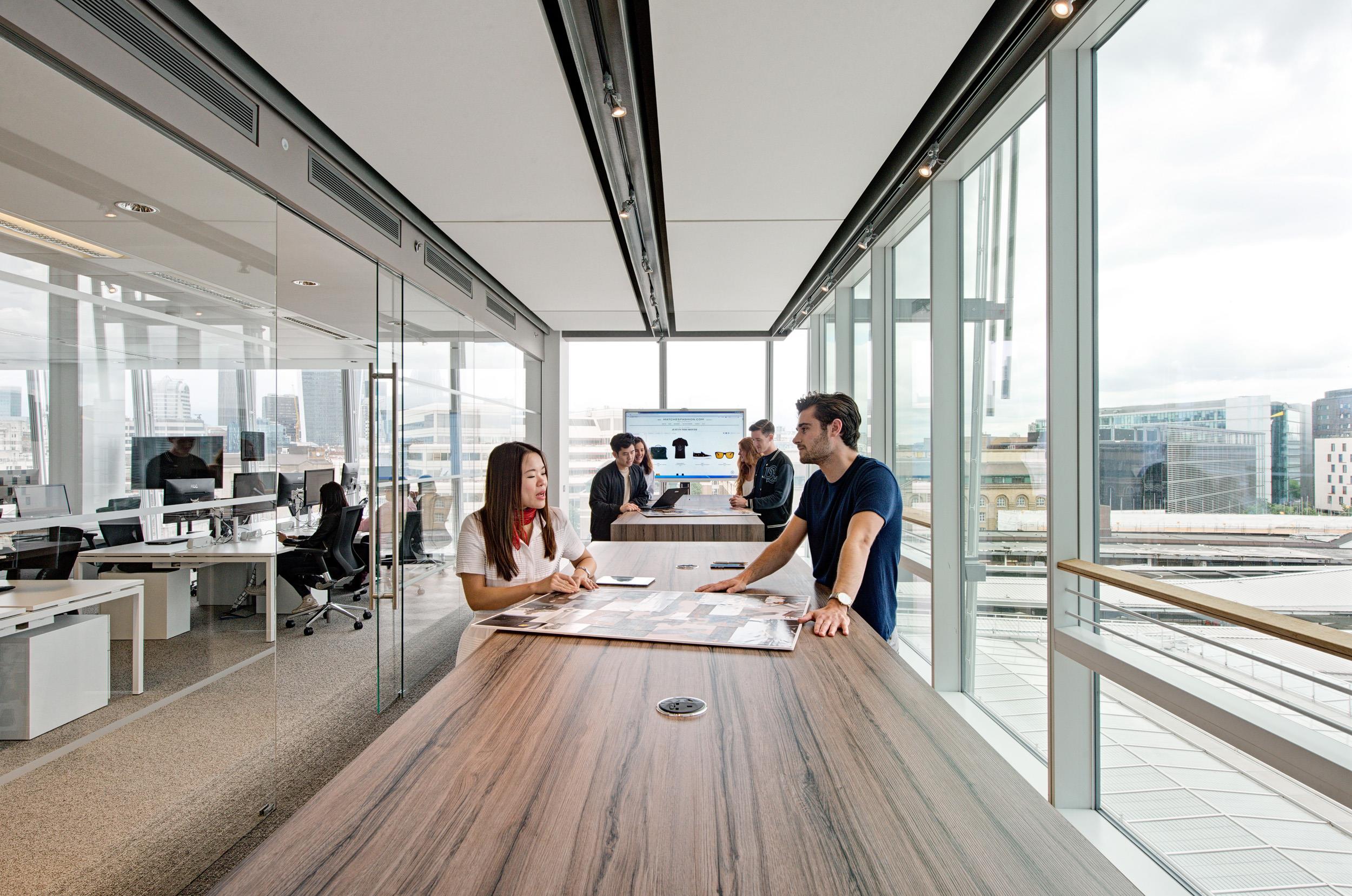 Collaborative work zone for Matches Fashion, with tall wood-effect tables and integrated power units set against a backdrop of full-height glazing and city views.