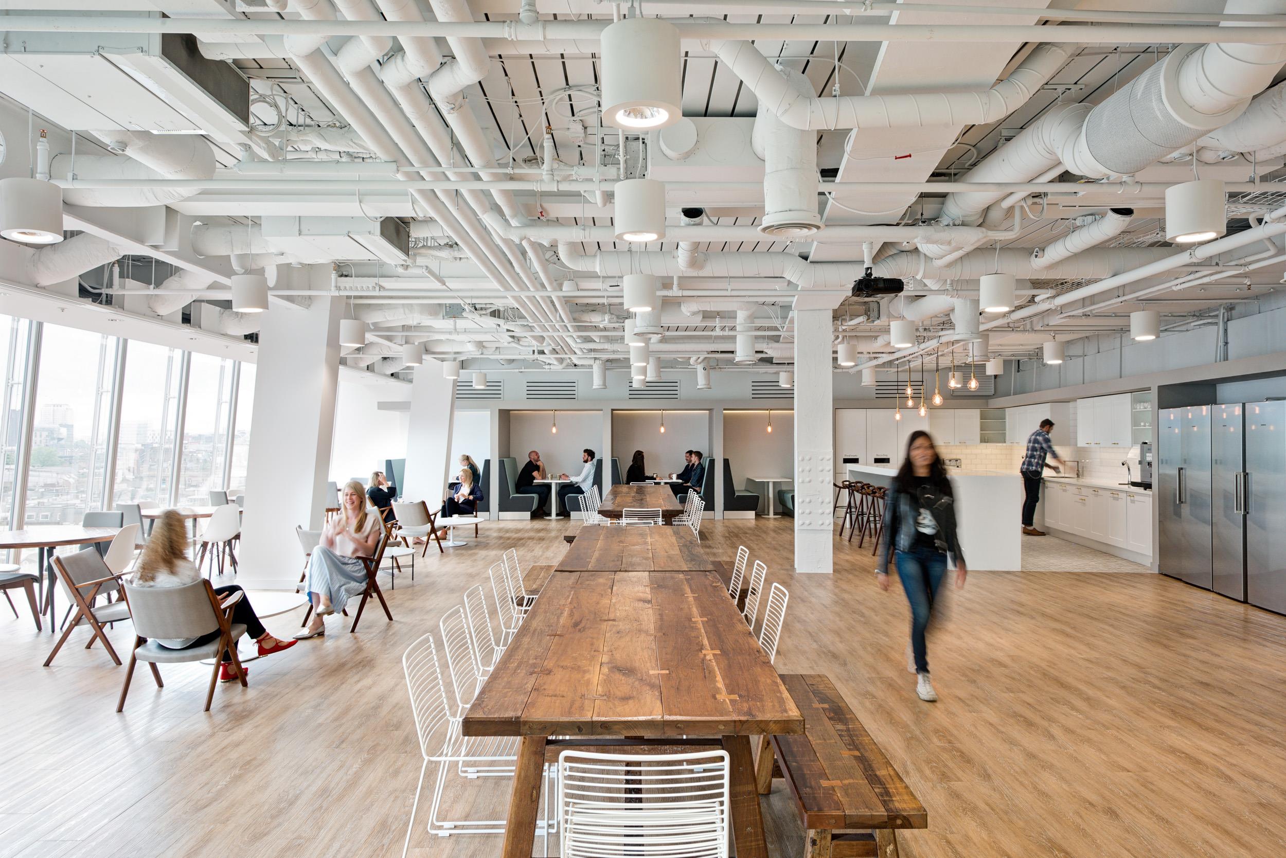 Canteen and breakout space for Matches Fashion, featuring long timber tables with white wire chairs, lounge seating, and kitchen facilities beneath an exposed ceiling.