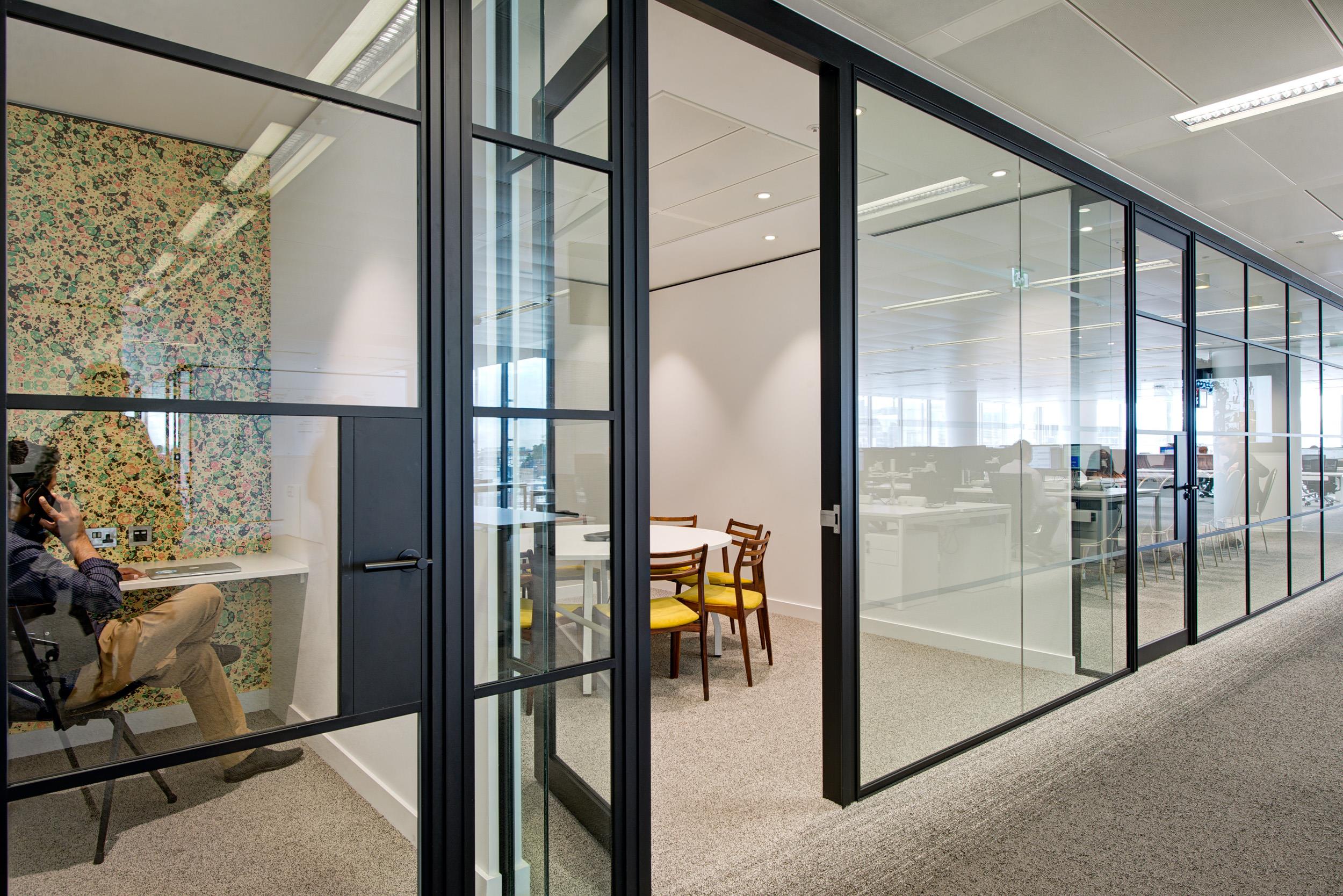 Private workspace at Matches Fashion, featuring a phone booth with floral wall covering and a glass-partitioned meeting room with timber chairs and yellow seat pads.