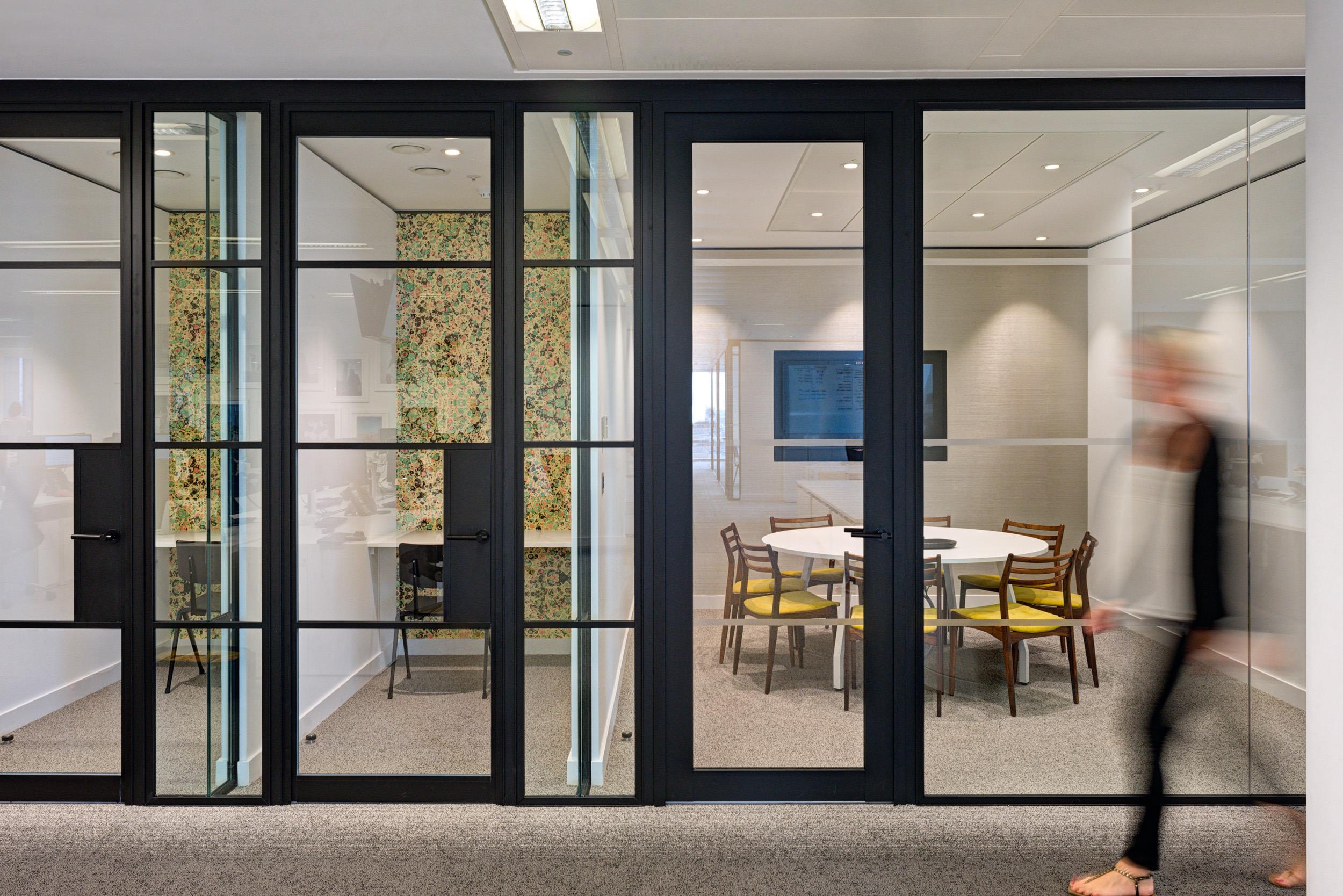 Glazed meeting rooms and phone booths at Matches Fashion, styled with floral wallpaper, dark-framed partitions, and yellow-upholstered chairs around a white table.
