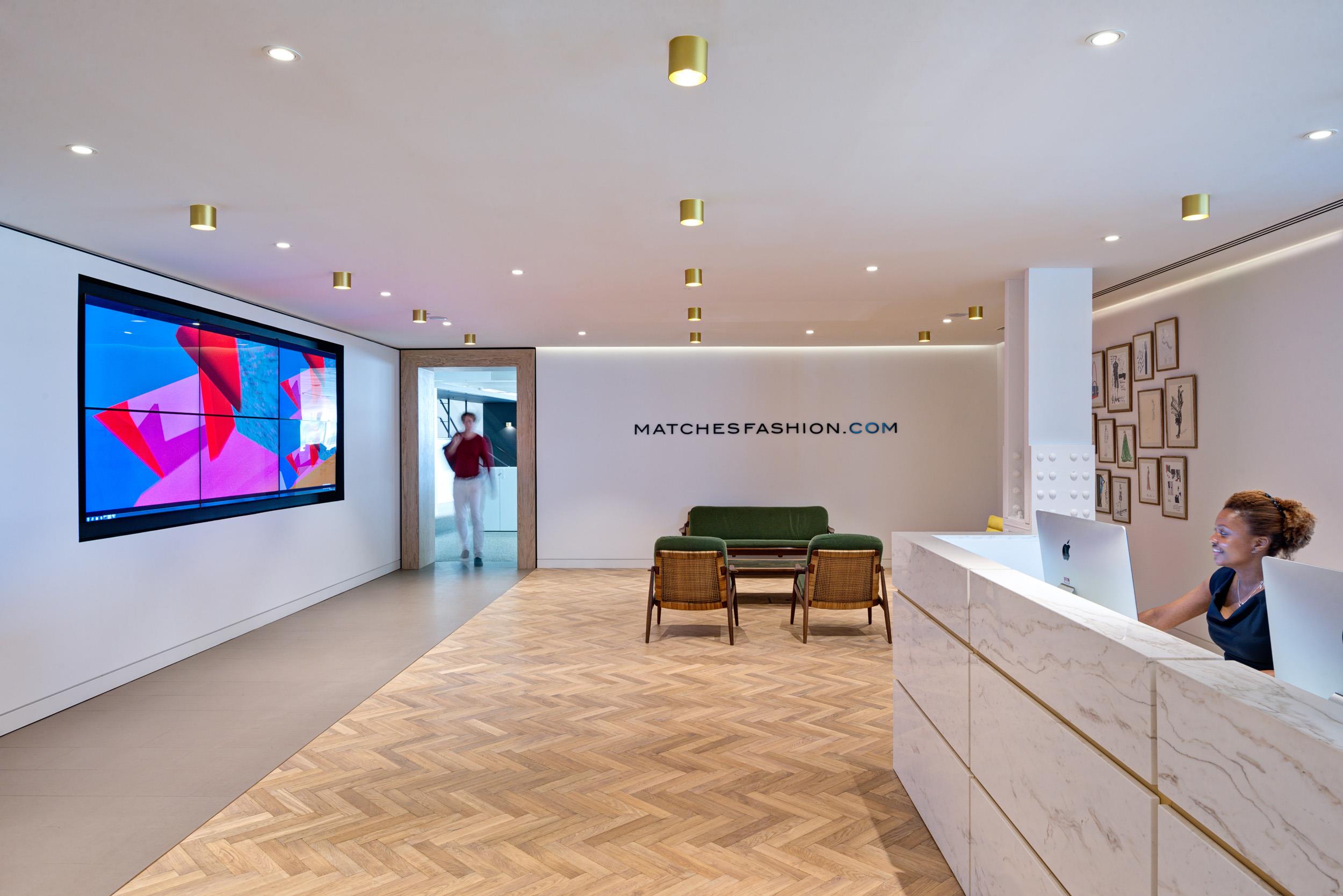 Reception area at Matches Fashion, showcasing a marble desk, green lounge seating with cane backs, a wall-mounted screen, and parquet flooring.