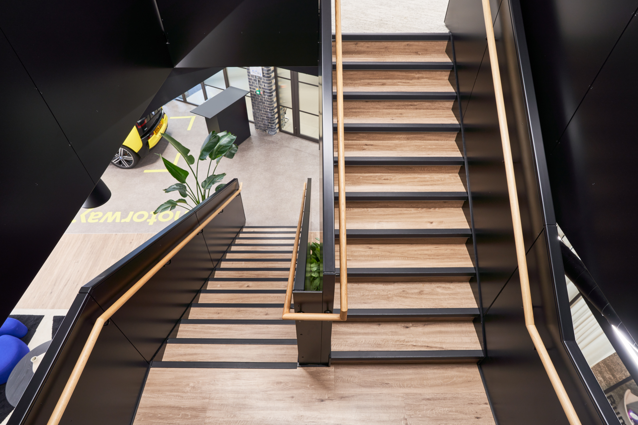 View down the central staircase at Motorway, revealing timber treads, black balustrades, and glimpses of the reception space and electric car display below.