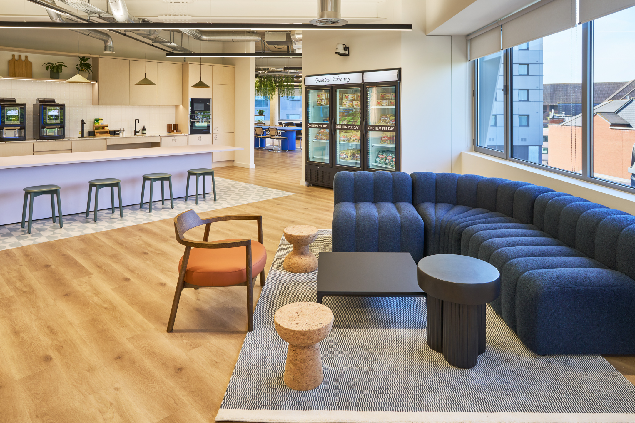Teapoint and breakout area at Nomad Foods, furnished with a curved navy sofa, cork stools, and a minimalist kitchenette designed for informal meetings and relaxation.