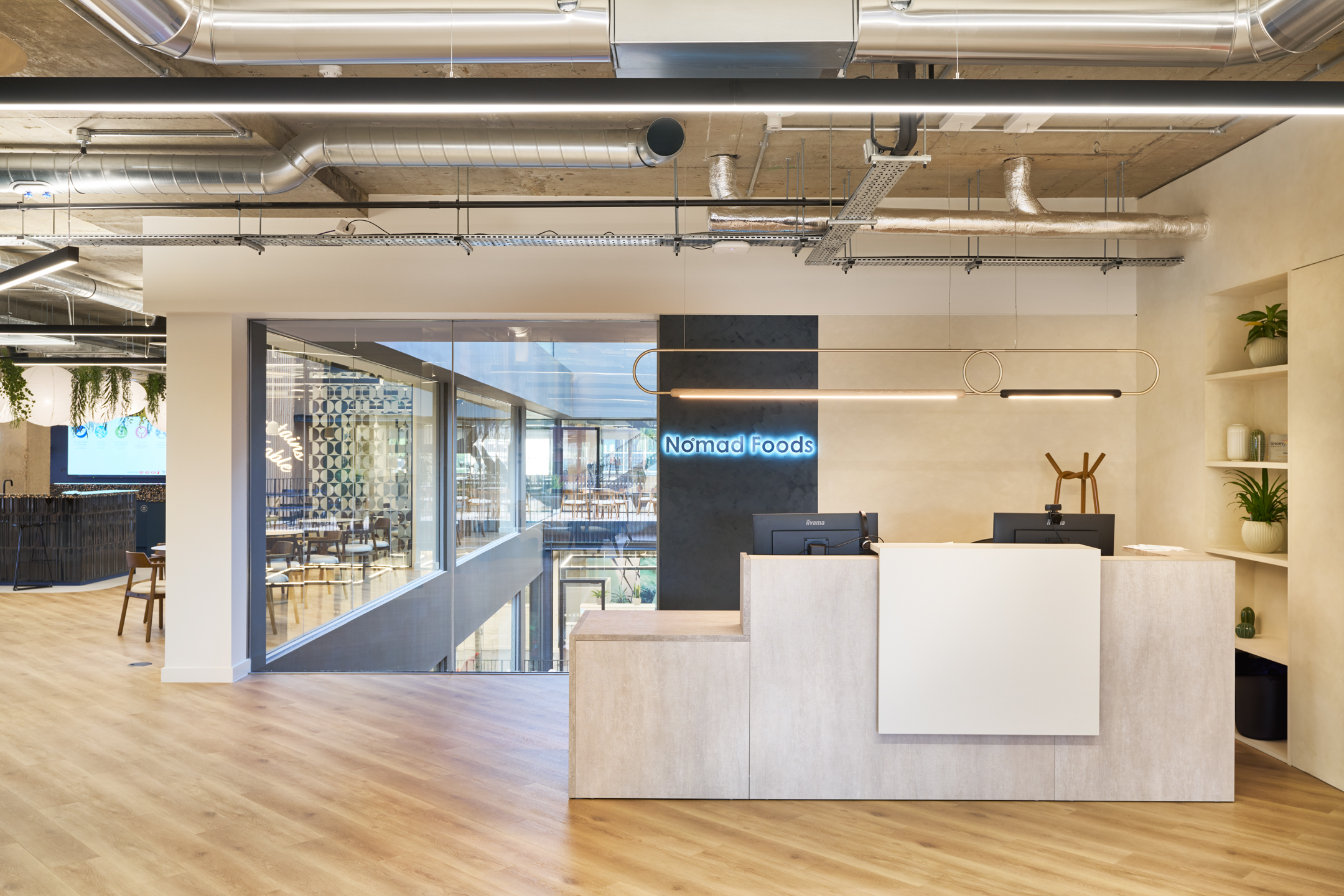 Reception area at Nomad Foods, featuring a clean-lined desk, open shelving with greenery, and a glowing brand sign set against an industrial-style ceiling.