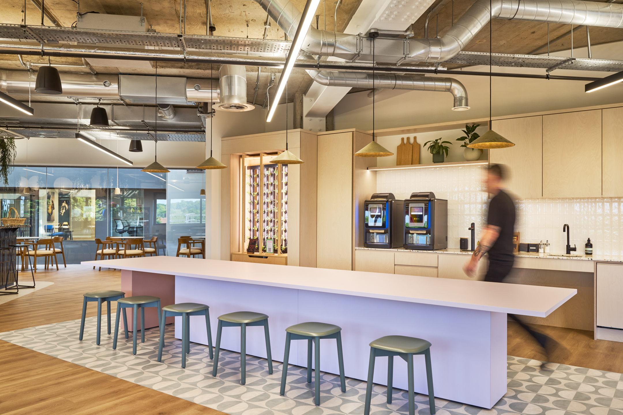 Light-filled teapoint at Nomad Foods with a long communal island, green stools, filtered water and coffee stations, and a hydroponic herb wall for a wellness-inspired touch.