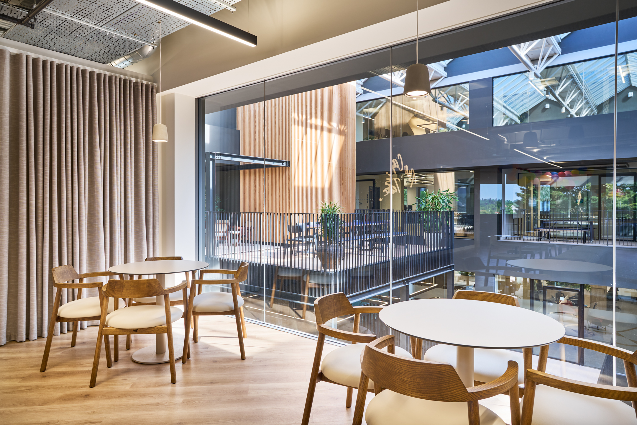 Informal dining space at Nomad Foods with round tables and timber-framed chairs, overlooking a central atrium with floor-to-ceiling glazing and views across the office courtyard.