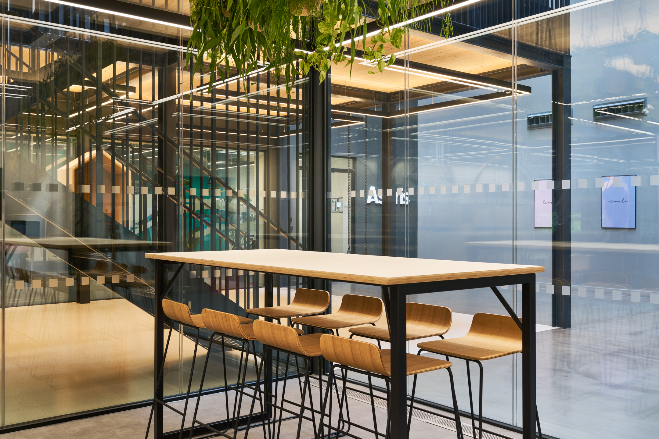 High table and bar stools in a glass-enclosed collaboration space at Nomad Foods, positioned beside an open staircase and enhanced with suspended greenery overhead.