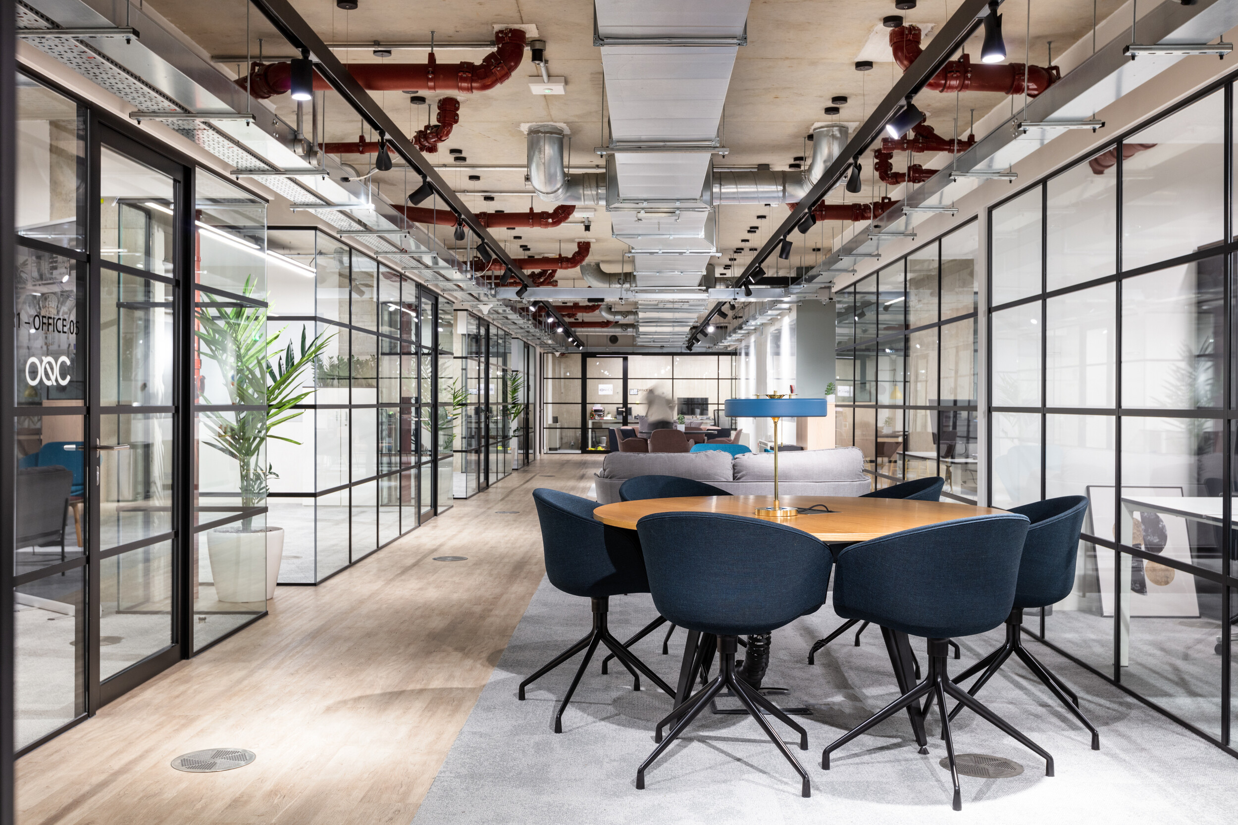OSE collaborative zone with blue upholstered chairs around a timber table, framed by industrial glazing and exposed ceiling services.