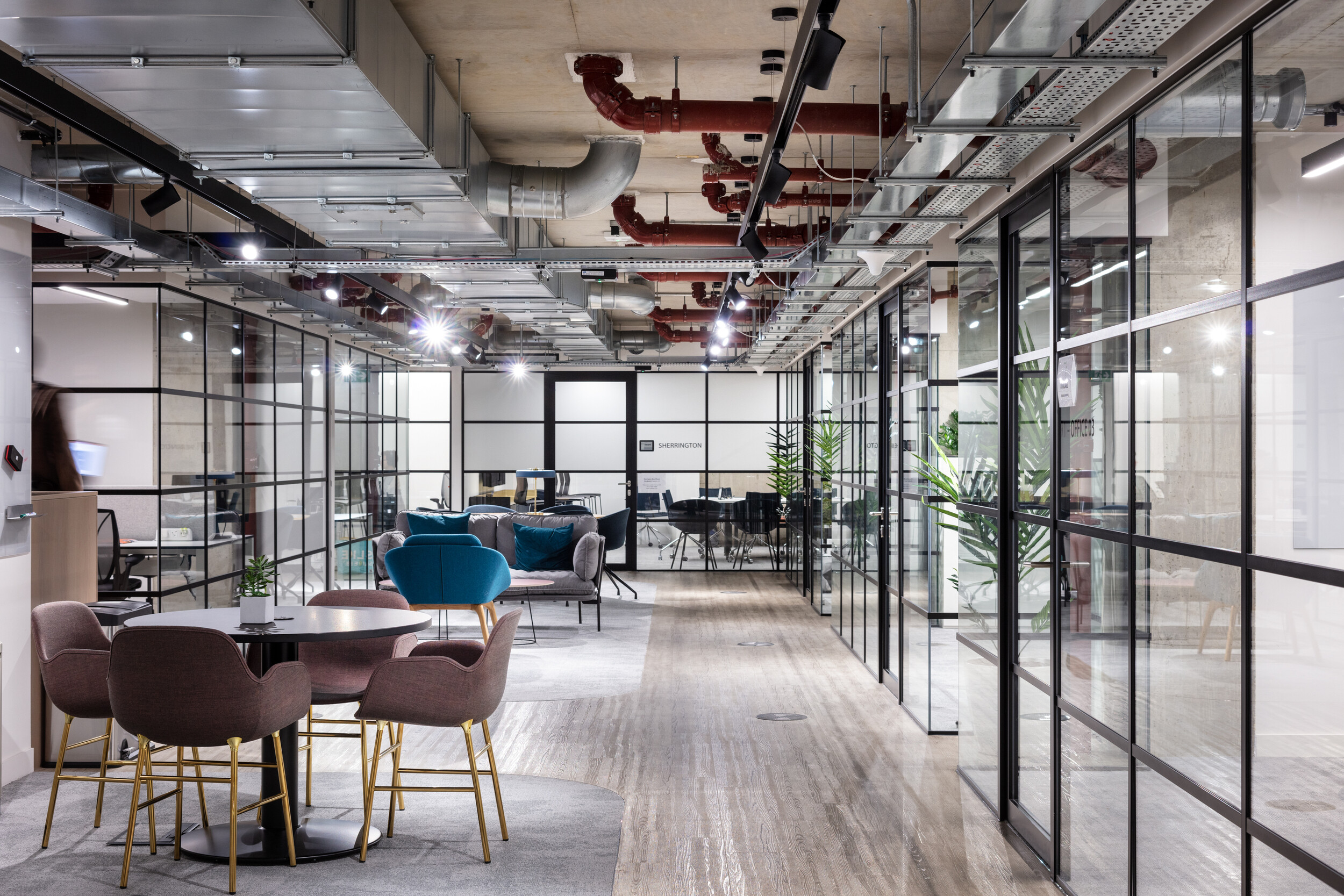 OSE workplace corridor with furnished breakout zones, including soft seating in teal and mauve, paired with gold-framed chairs and round tables.