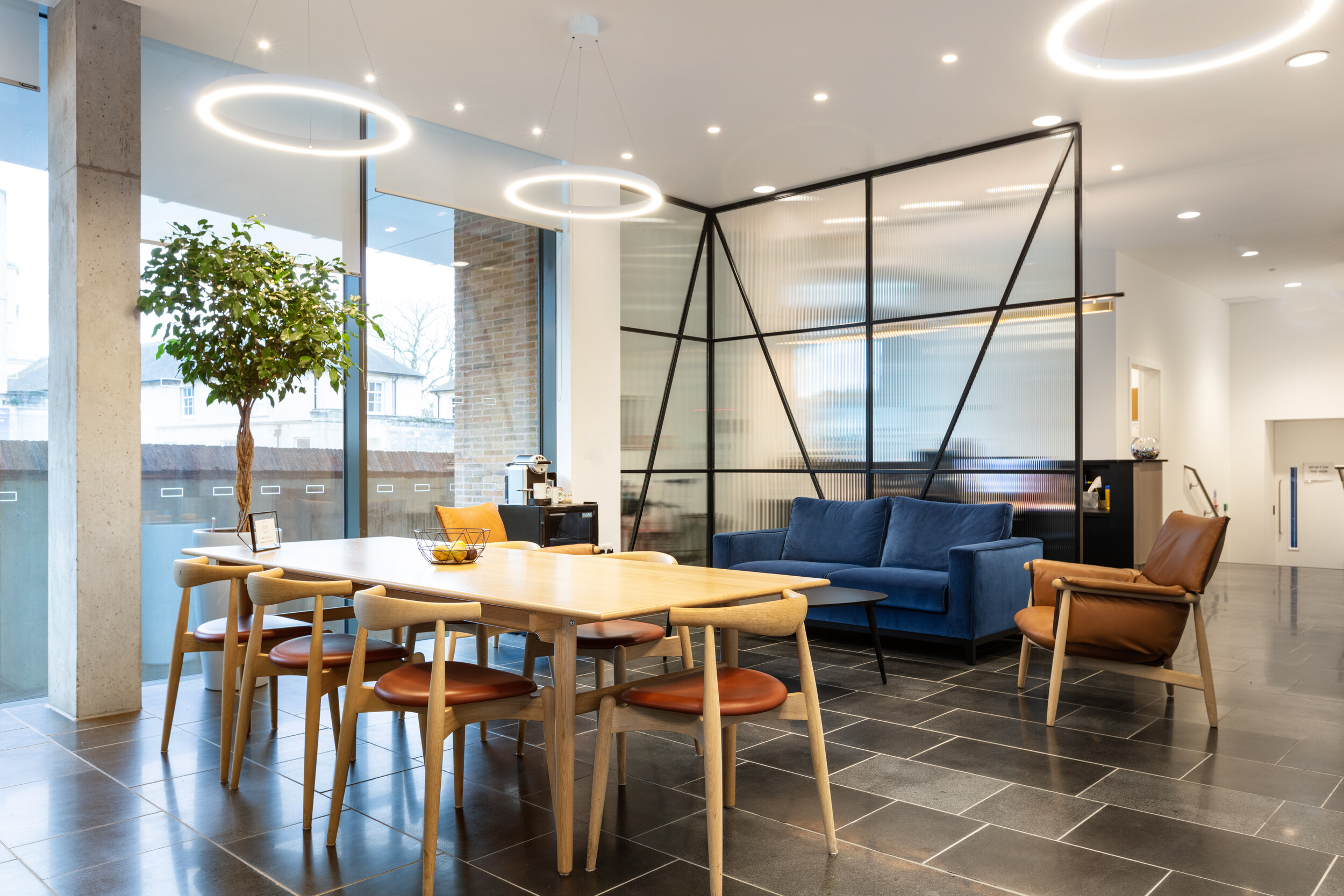 OSE informal meeting space featuring a timber dining table, tan leather and blue velvet seating, and contemporary lighting in a glazed corner zone.