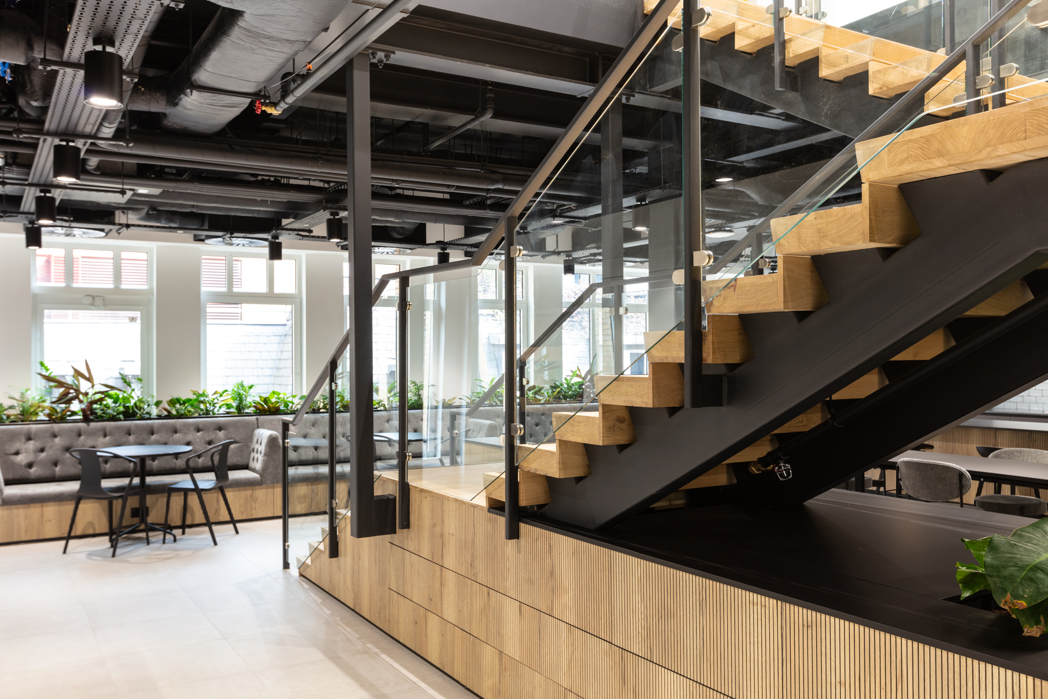 Furniture for Russell Reynolds Associates includes built-in banquette seating and round tables set beside a bold timber staircase and indoor planting.