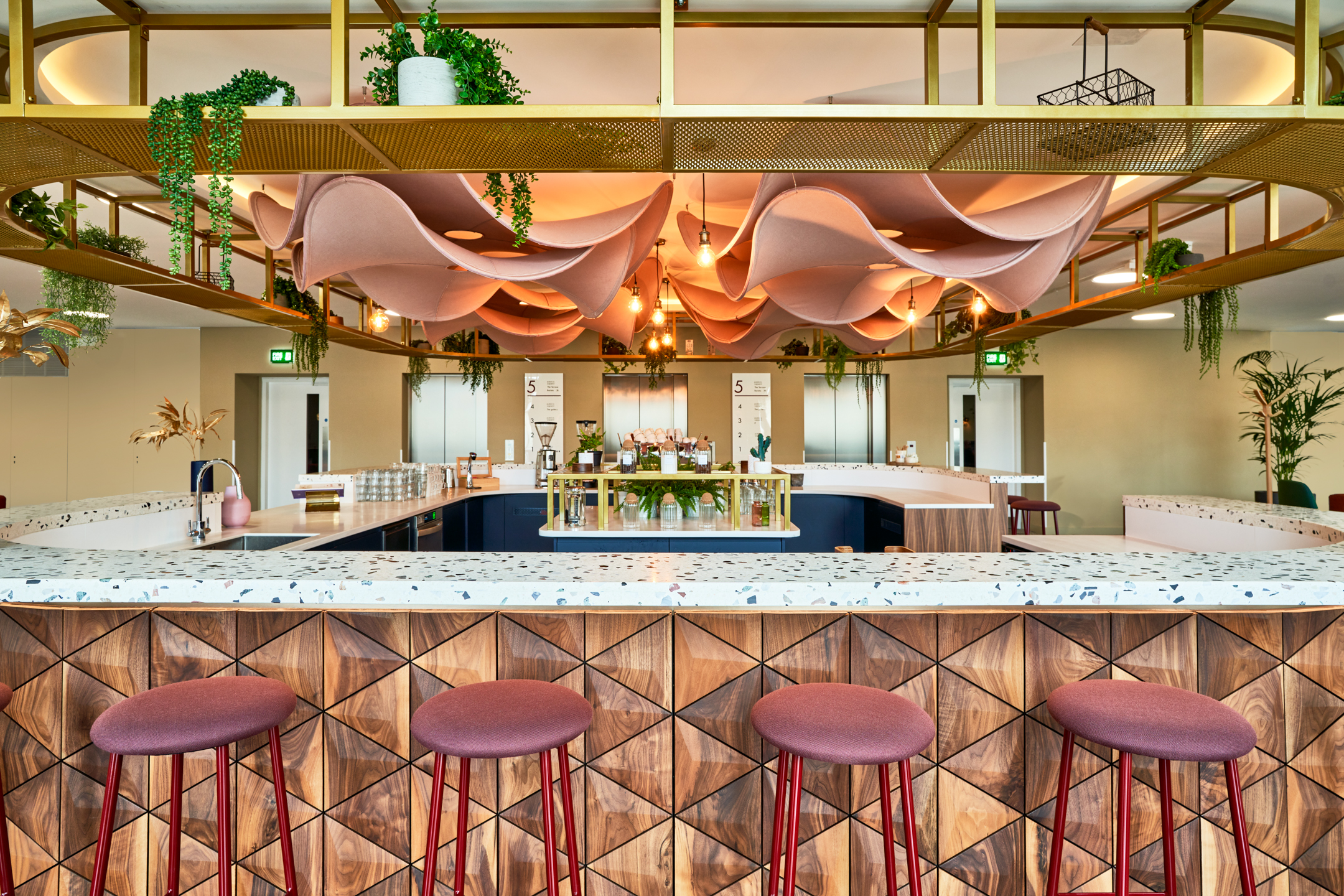 Central teapoint at Publicis Sapient furnished by Platfform, with terrazzo worktops, geometric timber panelling, burgundy bar stools and overhead greenery and acoustic petals.