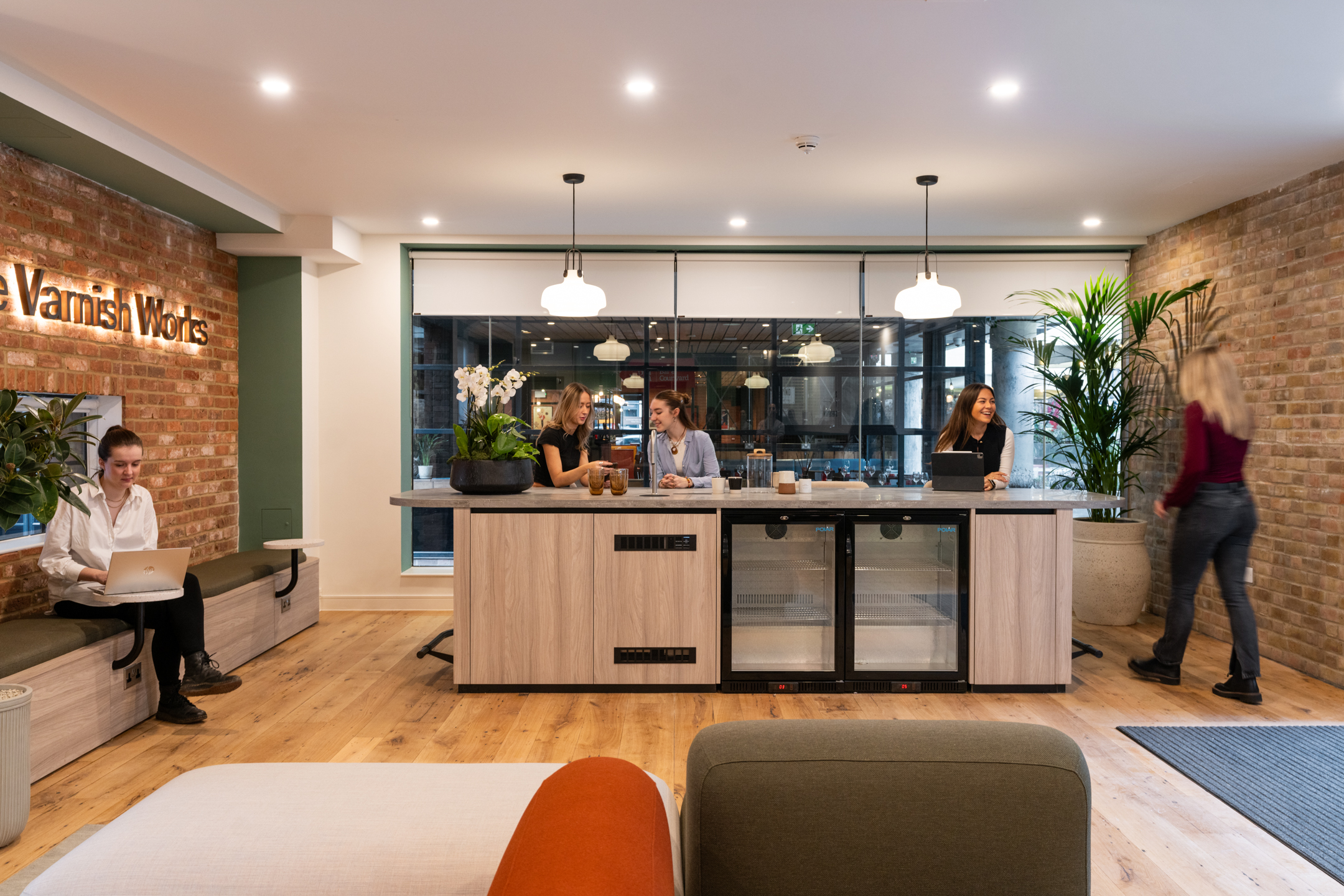 Light oak counter and soft bench seating at The Varnish Works styled with greenery and warm materials against exposed brick and industrial glazing