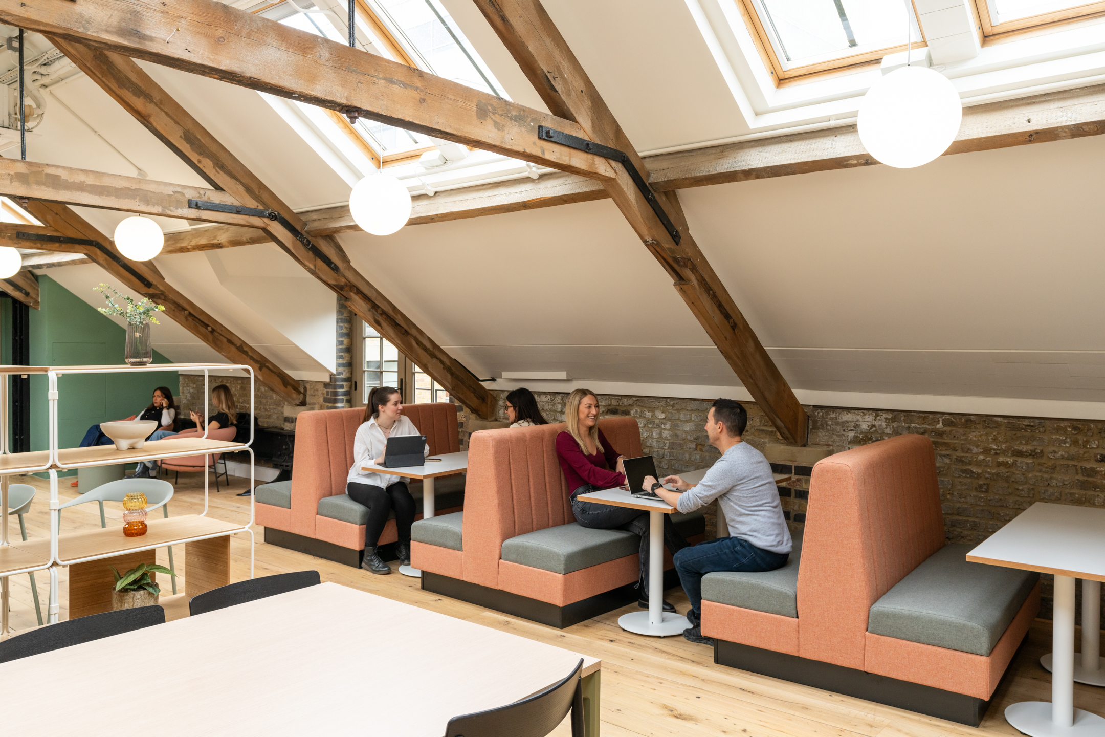 Bold booth seating in coral and sage lines the attic workspace at The Varnish Works, framed by exposed beams and soft light from roof windows
