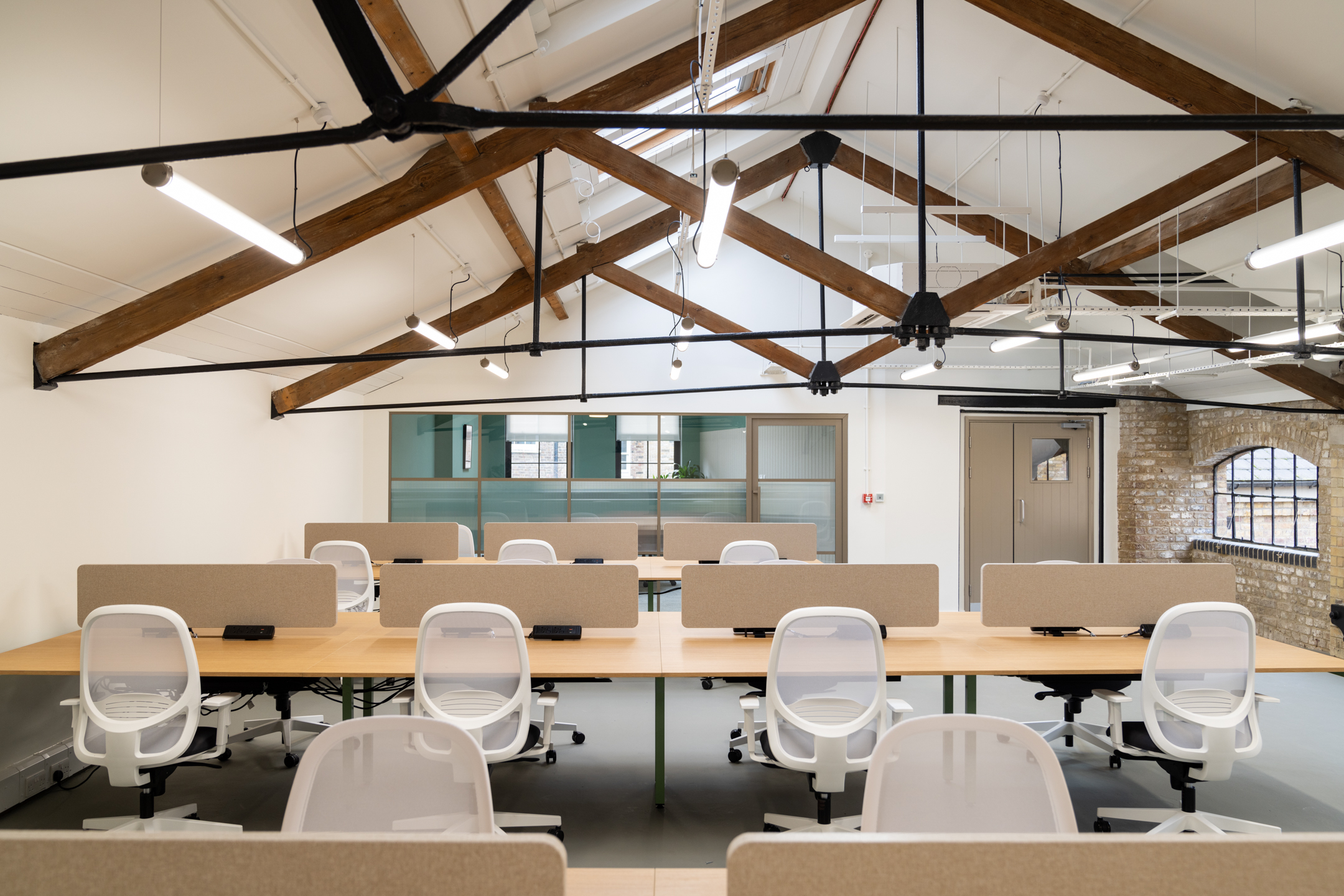 Desks with soft-backed screens and mesh chairs fill the vaulted studio at The Varnish Works, finished with timber beams and factory-style windows