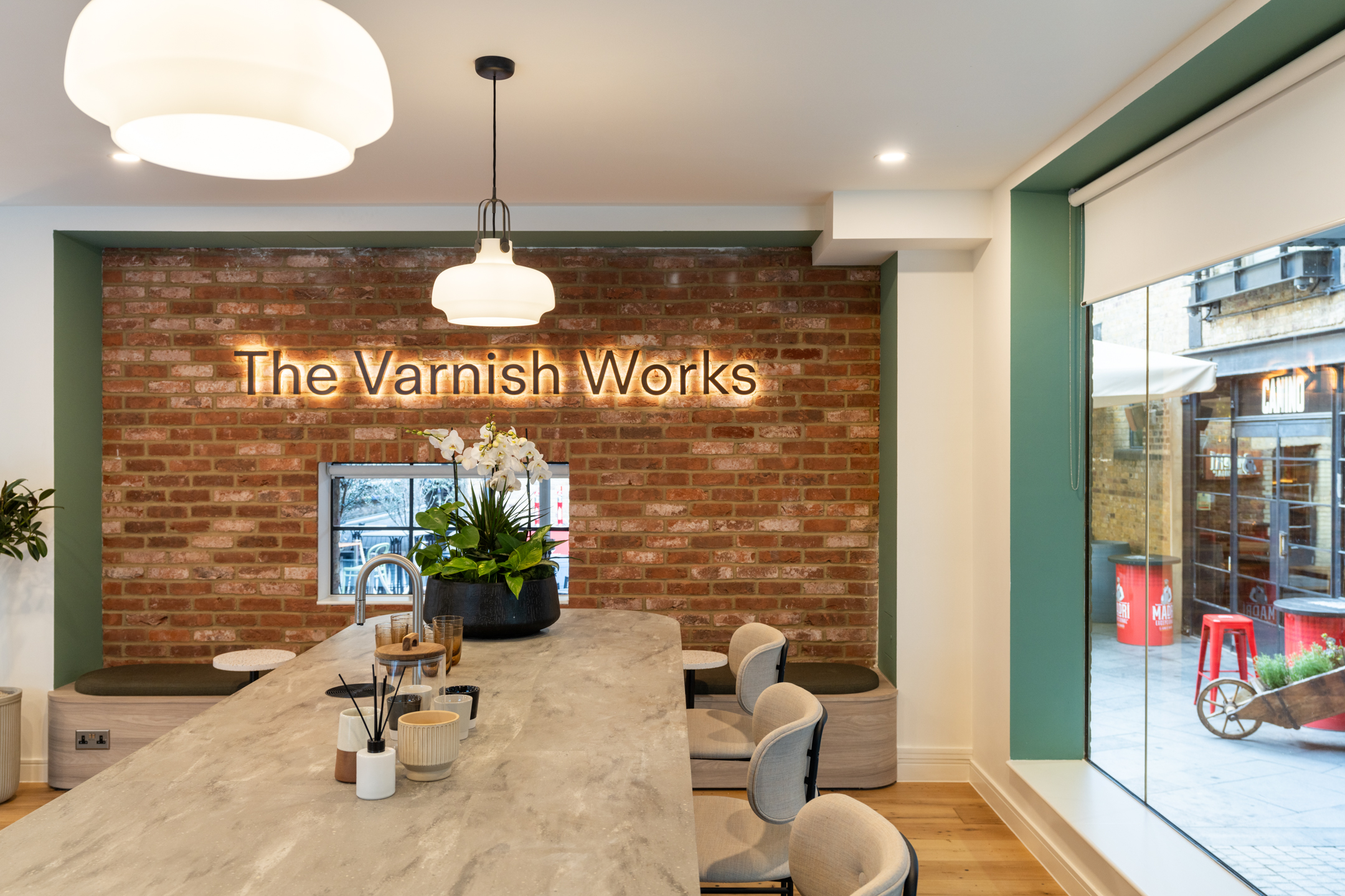 Bar stools and curved benches bring softness to the entrance at The Varnish Works, set against exposed brick and a glowing heritage-inspired sign