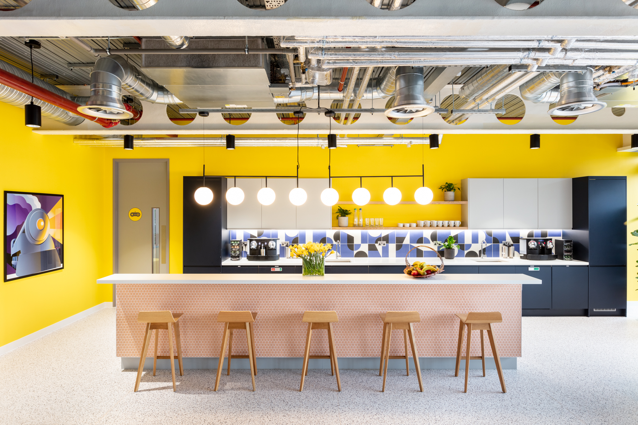 Travelfusion office kitchen with bespoke furniture from Platfform, featuring a bold yellow backdrop, pendant lighting and tailored bar stool seating.
