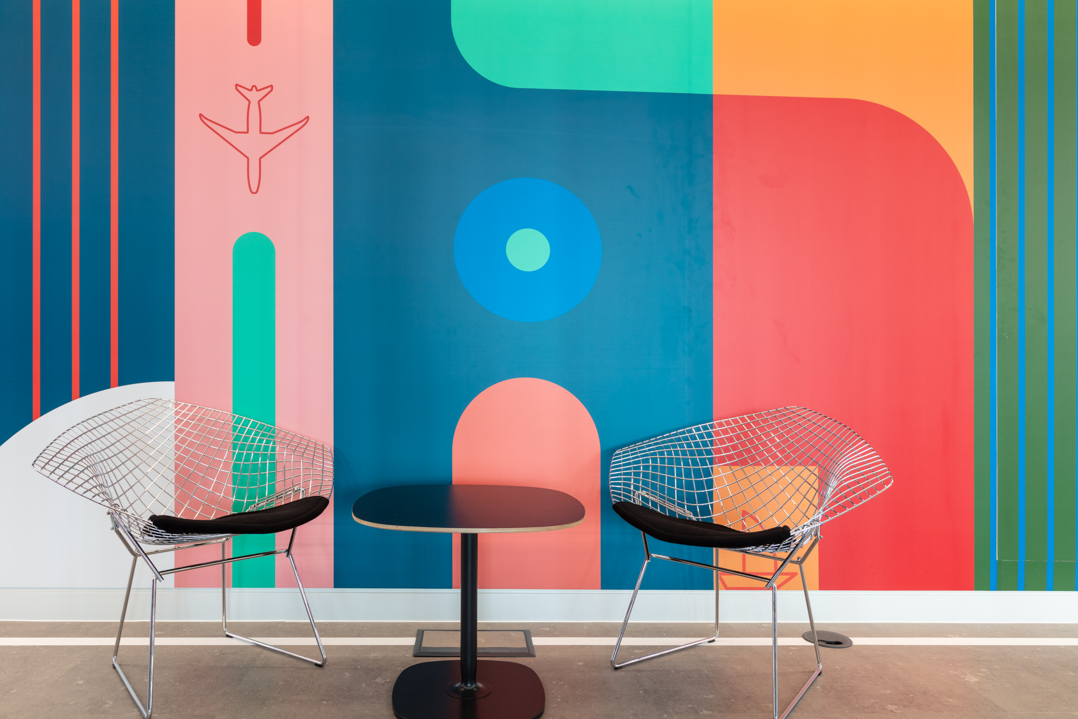 Travelfusion breakout nook with wire-frame lounge chairs and a compact café table by Platfform, set against a bold mural with aviation-inspired graphics.