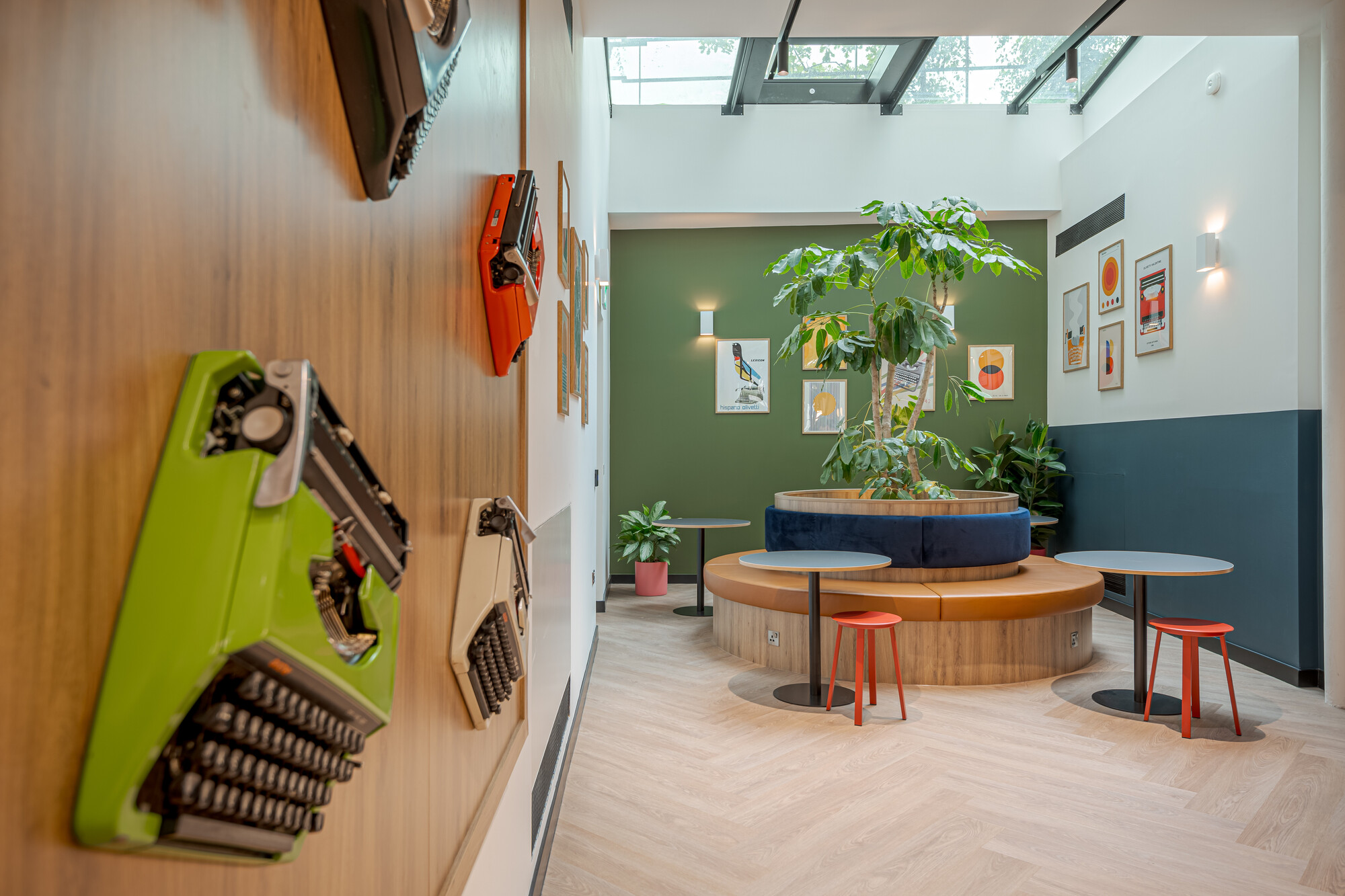 Work.Life Typewriter breakout area with vintage typewriter display and central banquette seating under a skylight, framed by green and timber tones.
