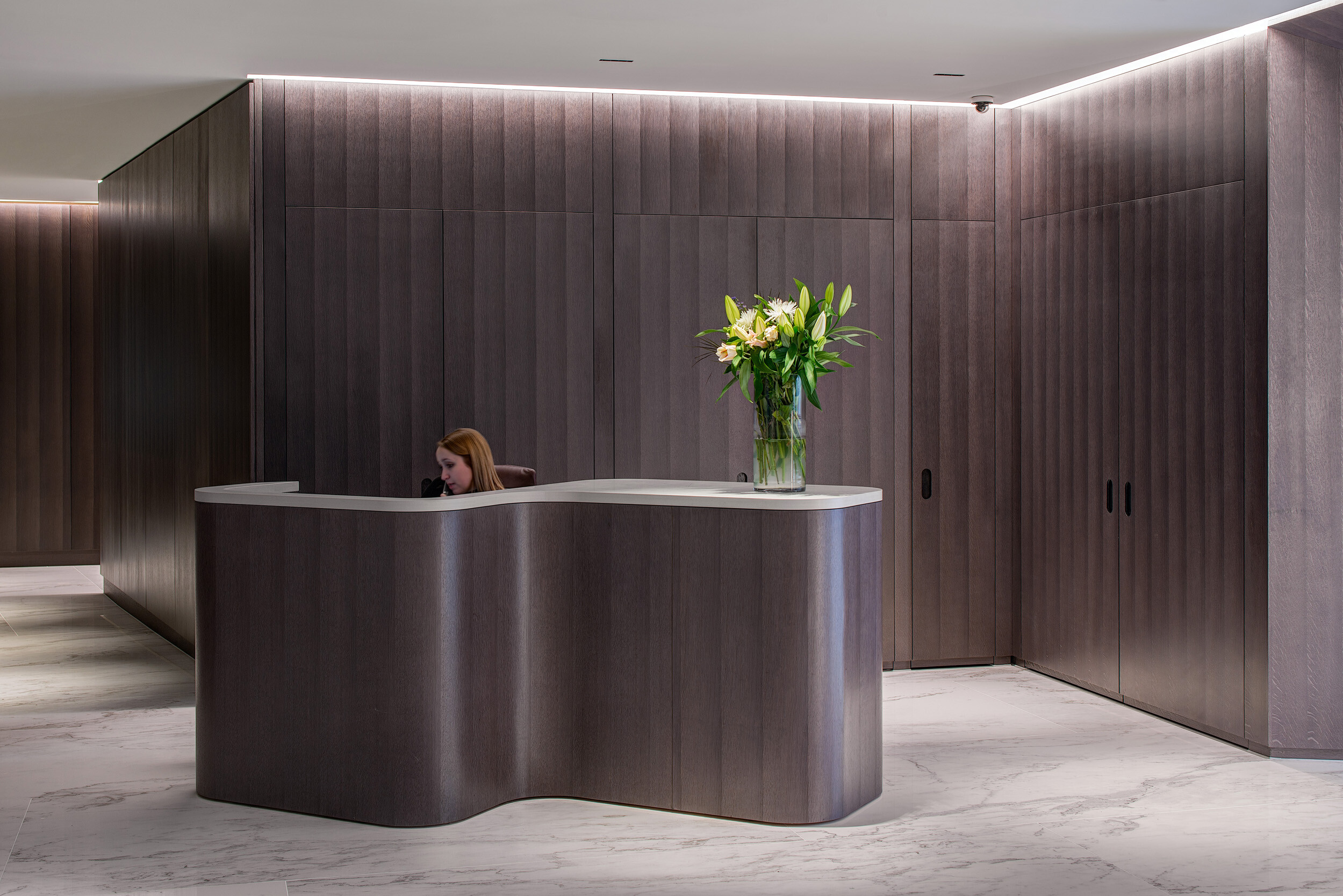 Reception desk at Vedanta’s London office with curved dark wood panelling and fresh flowers, reflecting the refined aesthetic of the workspace.