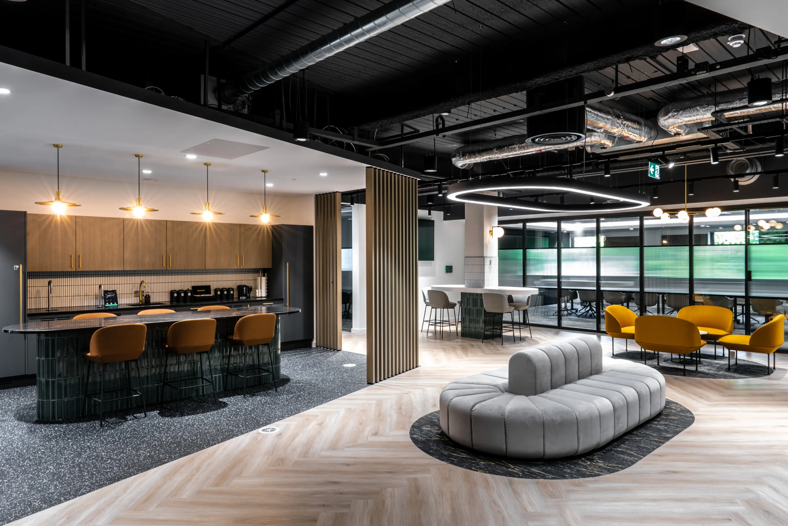 breakout space at Watches of Switzerland with bold yellow seating, curved grey sofa, and dark framed glazing across the back wall.