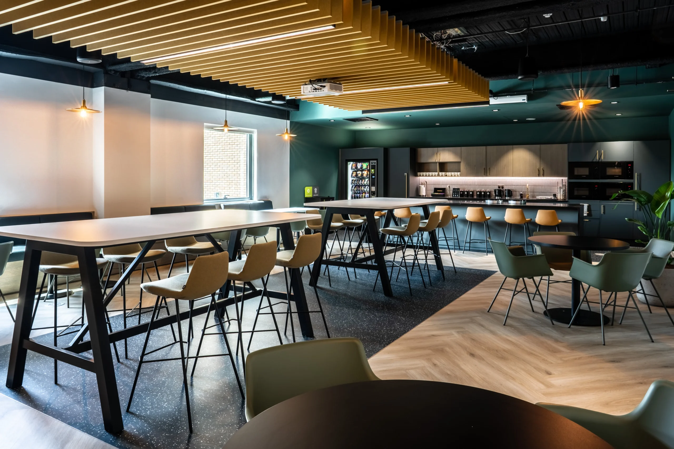 Larger teapoint at Watches of Switzerland with long shared tables, green and yellow seating, and a sleek kitchen set beneath golden ceiling fins