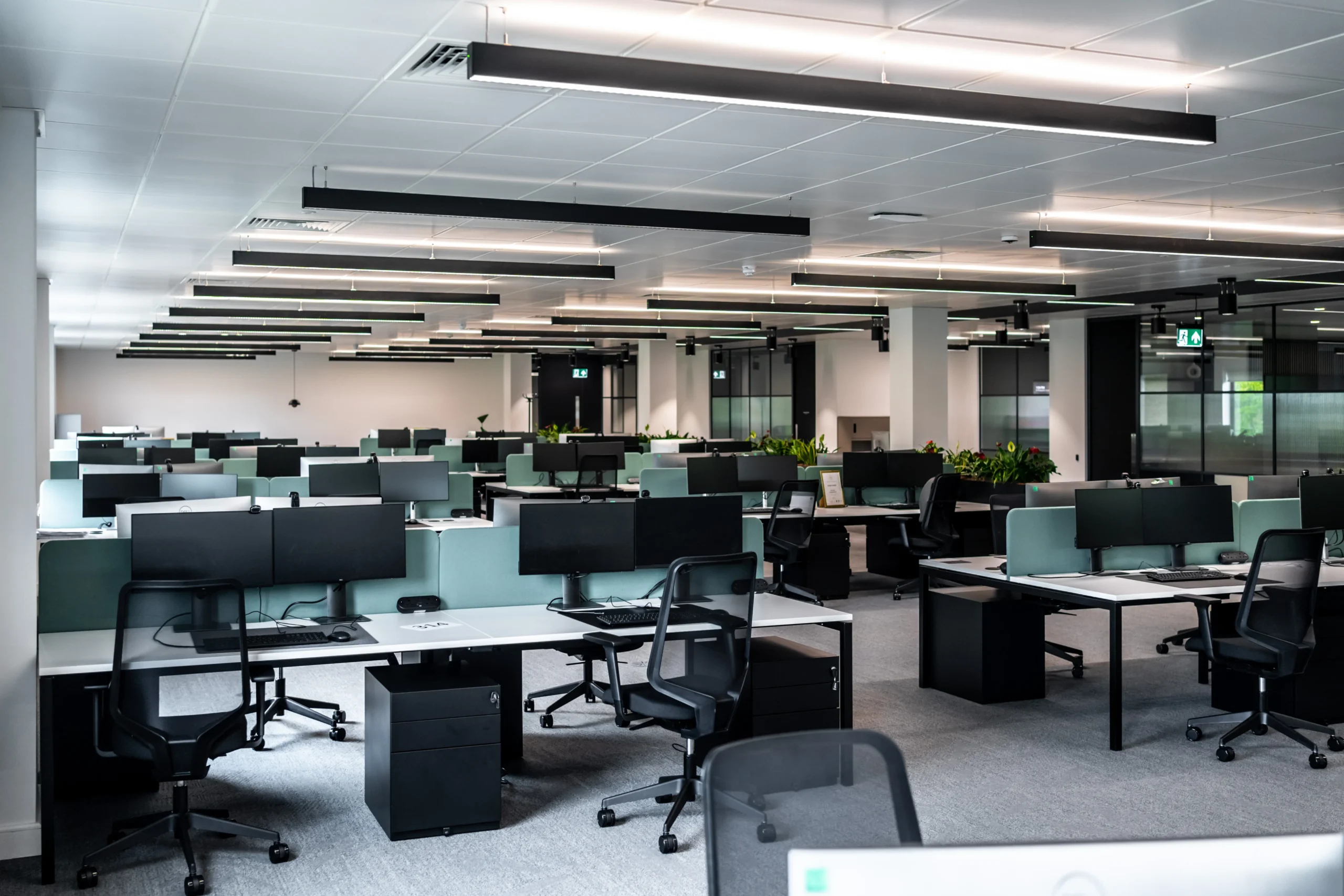 Open plan workspace at Watches of Switzerland with dual-monitor setups, green privacy screens, and black task chairs across neat desk clusters